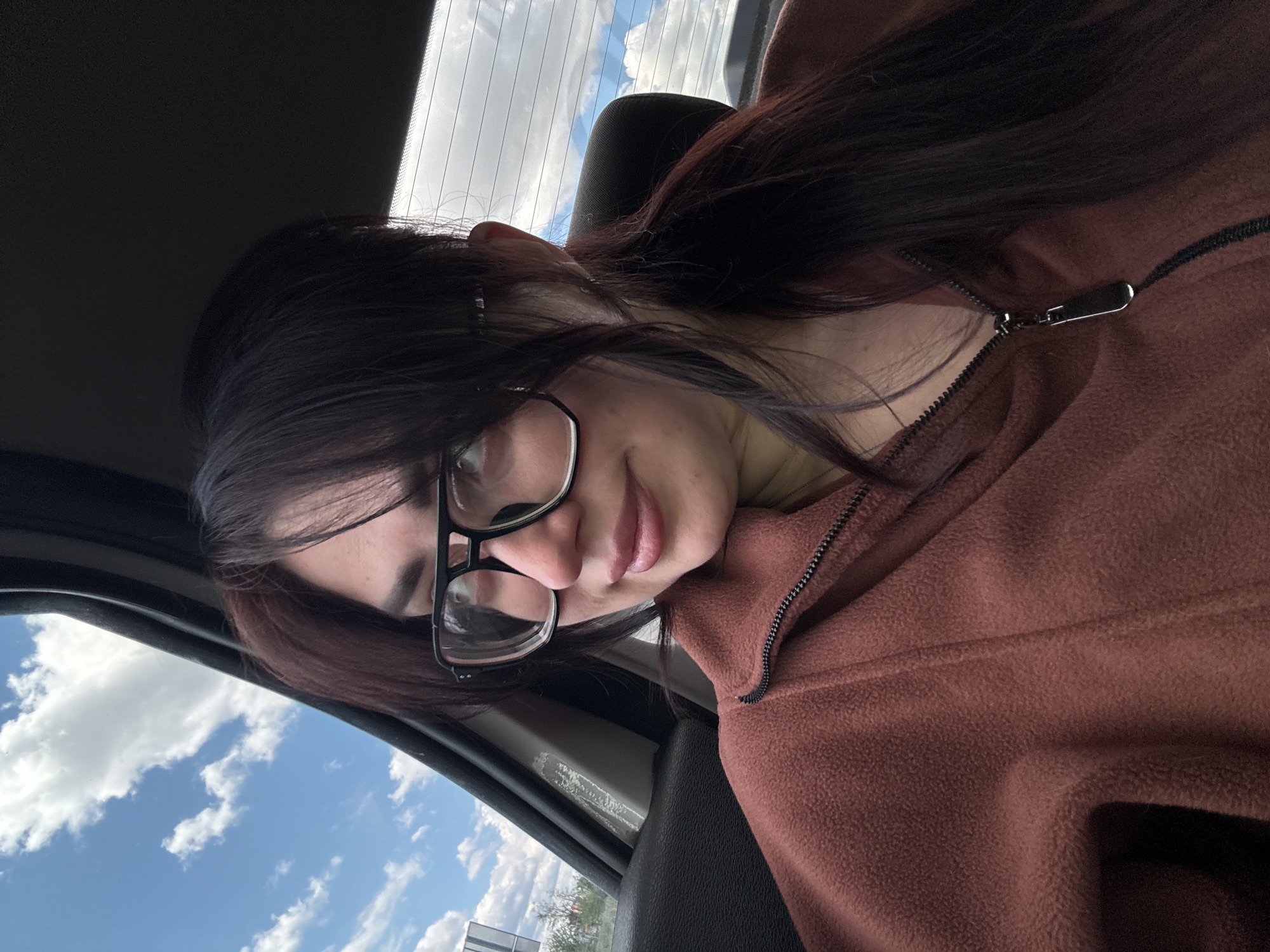 A woman is pictured in a close-up, selfie-style photograph, likely taken inside a car. She is wearing glasses with black frames and a brown fleece zip-up jacket. Her dark hair is parted and falls to one side. The background visible through the car window reveals a blue sky with white clouds. The location is inferred from the image content only.