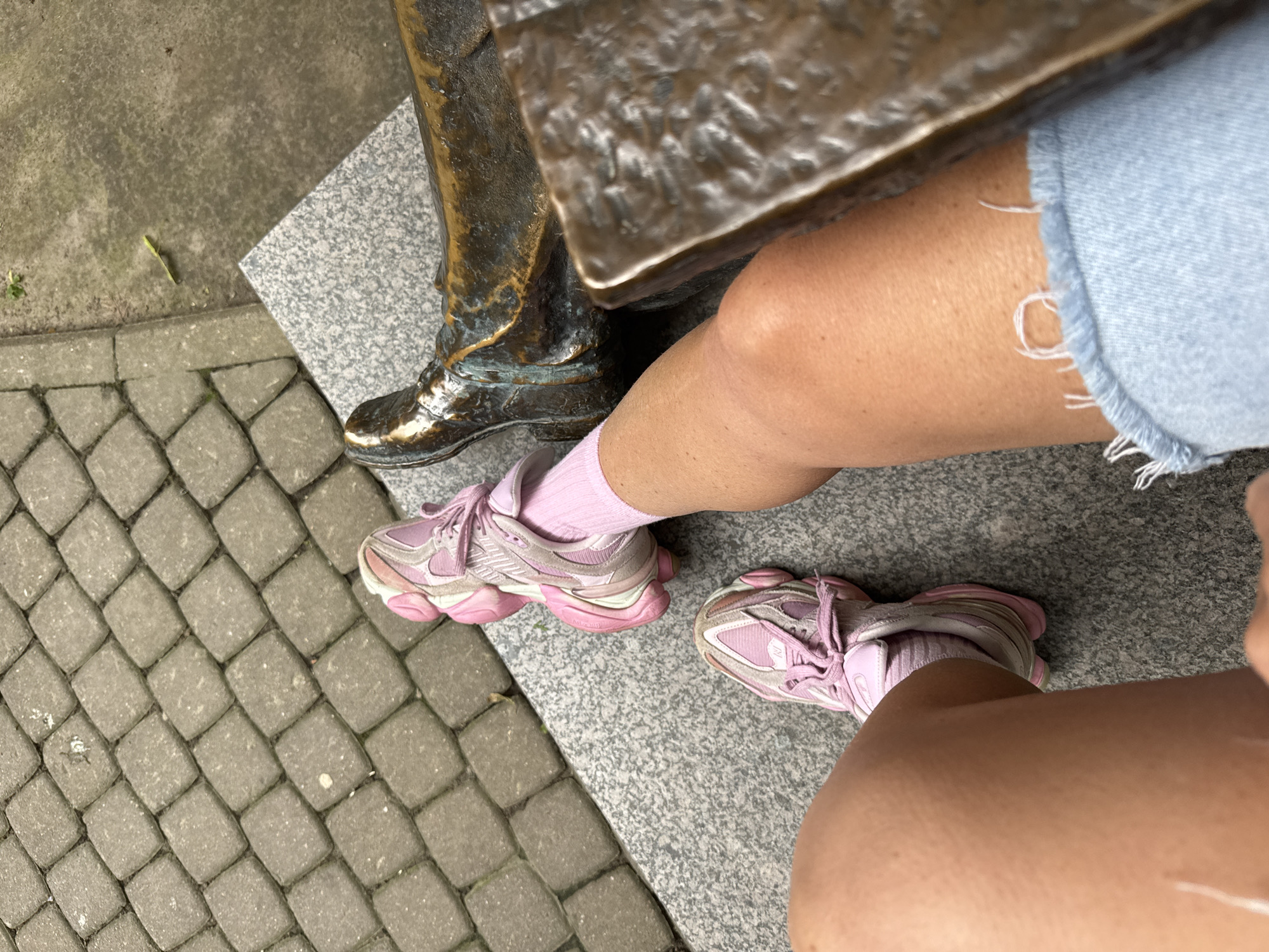 An overhead shot shows a person sitting on a granite bench next to a bronze statue of a person's legs, with a cobblestone pathway behind them. The person is wearing light blue denim shorts, pink socks, and pink athletic shoes. The statue's legs are clad in bronze boots, and the person's visible leg is tanned. The cobblestone pathway is laid in a basketweave pattern. The scene appears to be outdoors, possibly in a park or public square in Saint Petersburg, Russia.