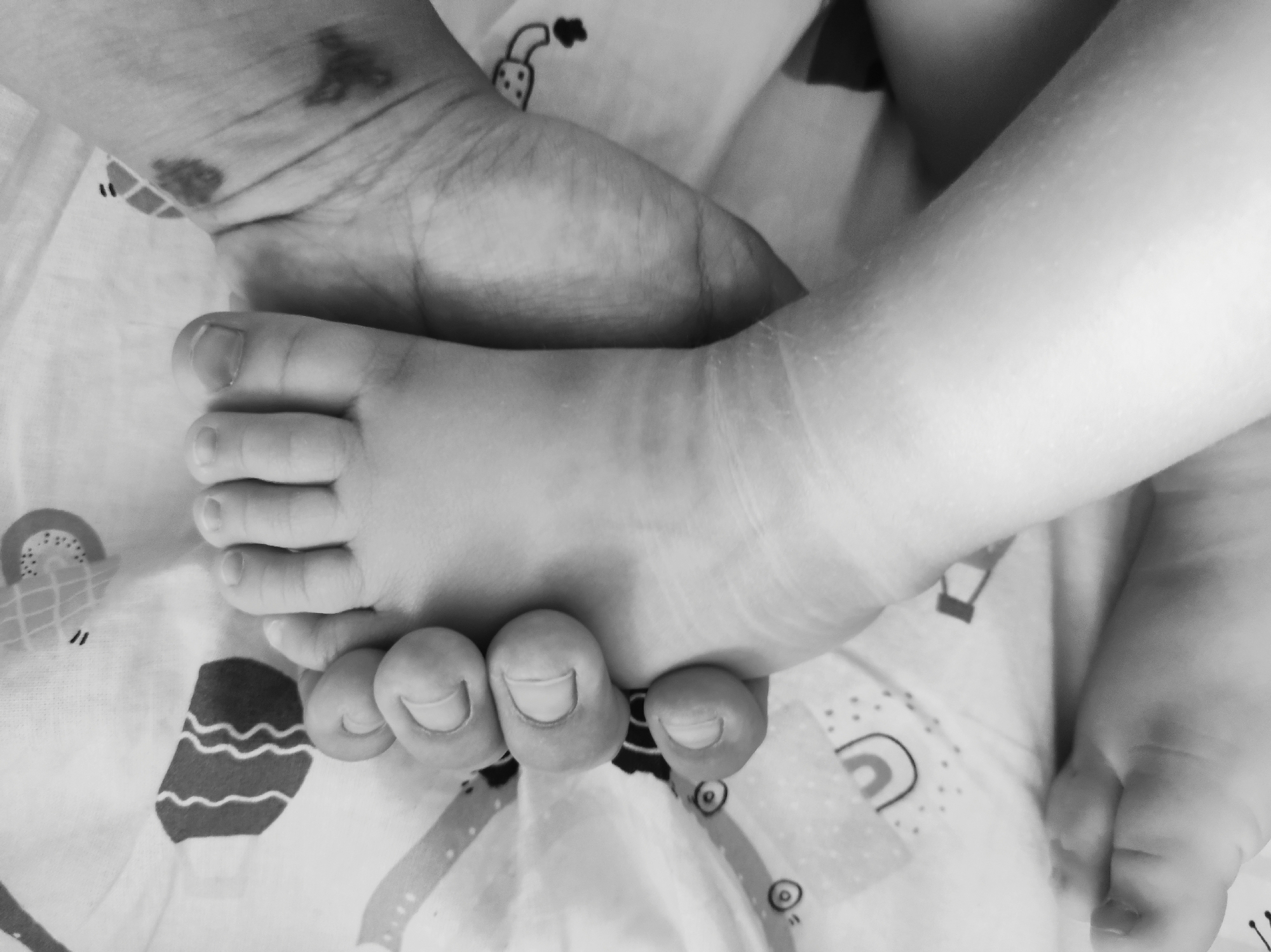 This black and white, overhead close-up shot features two human feet, likely an adult's and a baby's, resting on a patterned fabric. The adult's hand is visible in the upper left, with a mark or birthmark on the palm. The baby's foot is placed on top of the adult's foot, with its toes clearly visible. The fabric beneath has a repeating pattern of simple illustrations, including what appear to be hearts, rainbows, and other childlike motifs. The lighting is soft and diffused, creating a gentle mood. The scene is intimate and suggests a moment of connection between the two individuals. No text is visible in the image. The location context of Chaikovsky, Russia, does not provide any specific visual cues within the image itself.