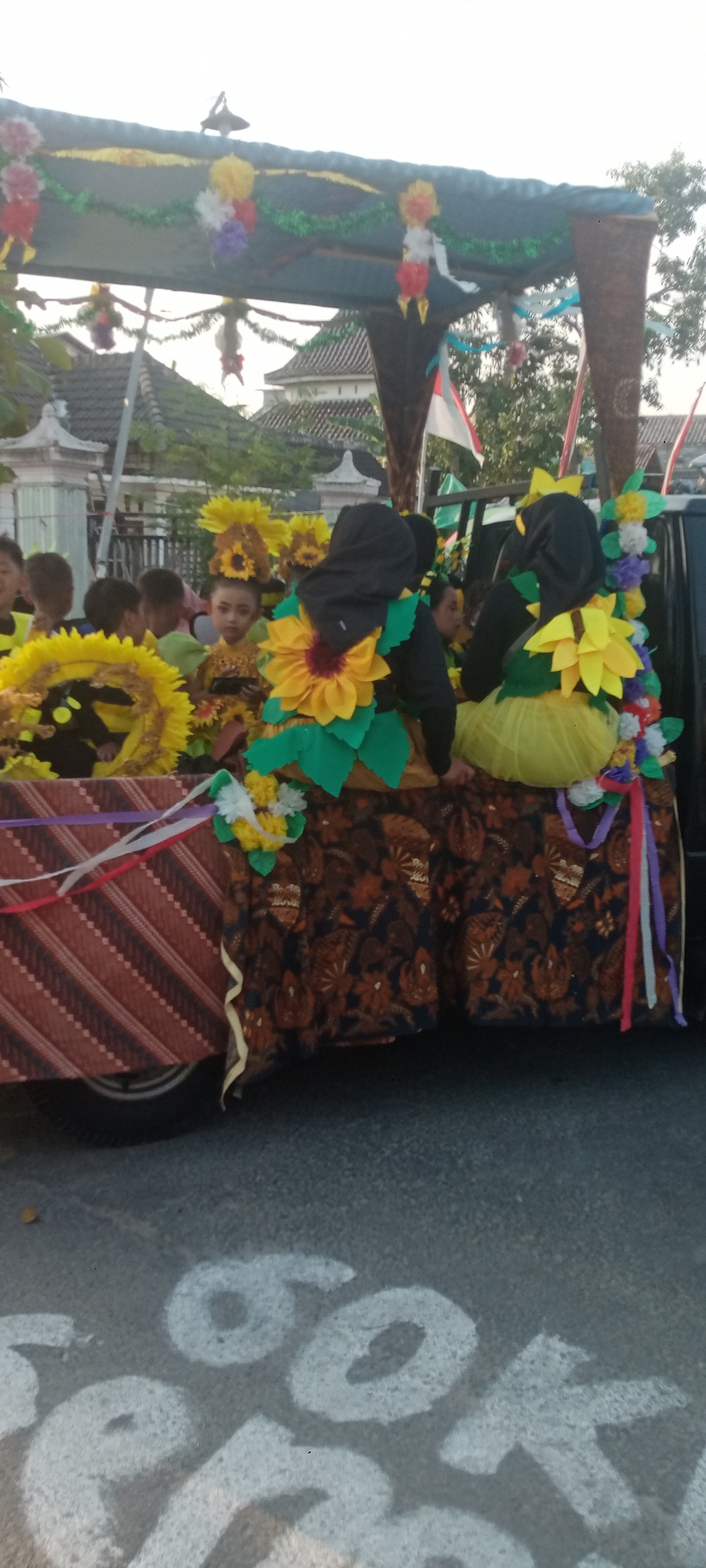 In Soko, Indonesia, a parade float is adorned with sunflower motifs and colorful decorations. Several children, dressed in costumes featuring sunflower elements and headwear, are seated on the float. The float itself is decorated with patterned fabrics and ribbons. In the foreground, large white chalk markings are visible on the asphalt road, partially obscured by the float. The background shows buildings and trees under a bright sky.