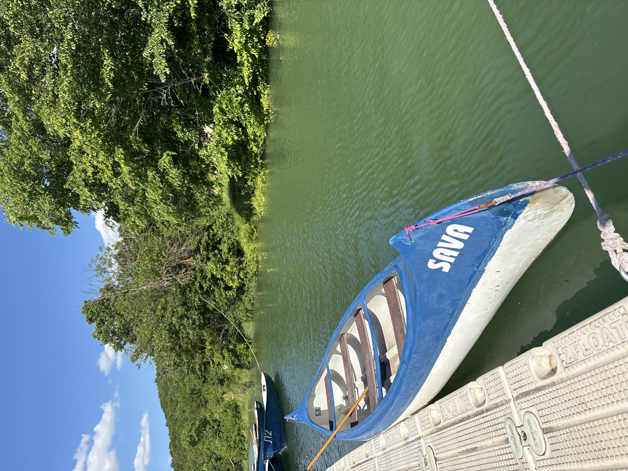 This image, captured in Sukko, Russia, presents a tranquil scene centered around a body of water, likely a river or lake, surrounded by lush natural vegetation under a bright sky.
In the foreground, a blue and white canoe, named "SAVA" in white capital letters on its stern, is moored alongside a light-colored, textured floating dock. The canoe is equipped with several wooden seating slats and a wooden paddle. Pink and purple ropes secure the canoe to the dock. The dock itself, made of plastic modules, has the word "2FLOAT" embossed repeatedly on its surface, indicating its purpose.
The water is a vibrant green color, reflecting the surrounding foliage and showing gentle ripples across its surface. To the left, another blue canoe, marked with "П2" (P2) on its stern, is partially visible, suggesting a line of rental boats. Beyond the canoes, the shoreline is densely covered with an abundance of green trees and bushes, indicating a rich, natural environment. The background features a clear, bright blue sky dotted with scattered white clouds, suggesting sunny weather.
The scene depicts an outdoor recreational setting, likely on a warm, sunny day. No people are visible, but the presence of the canoes and dock clearly points to boating or canoeing as the primary activity. The bright sunlight creates clear shadows and highlights the textures of the water and vegetation.