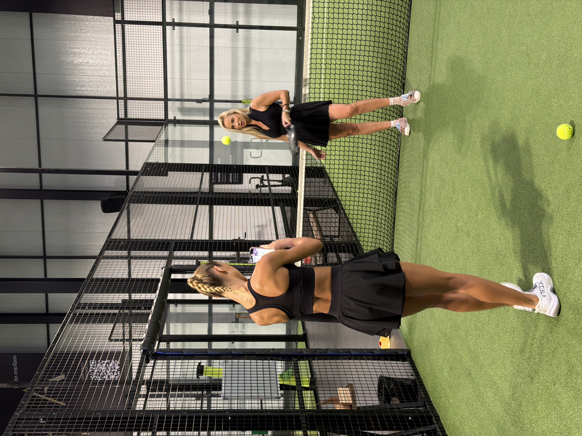 Two women are playing padel tennis on an indoor court. One woman, in the foreground, is facing away from the camera, with her back to the net, and appears to be retrieving a ball. She is wearing a black sports bra and a black tennis skirt, with white tennis shoes. Her hair is styled in a braid. The other woman is in the background, facing the net, and appears to be serving or hitting a shot. She is wearing a black tennis outfit and white tennis shoes with pink accents. A yellow padel ball is visible in the air between the two women. The court is made of artificial turf and is enclosed by glass walls and metal mesh. The ceiling is high with industrial lighting. The setting suggests an indoor sports facility. There is no indication of the time of day or weather, as the scene is indoors. Some visible text includes "XCROSS" and possibly brand names on the women's clothing and shoes. There are also Thai characters visible on the wall in the background, which aligns with the provided location context of Choeng Thale, Thailand. The atmosphere appears energetic and competitive, as is typical of a sports match.