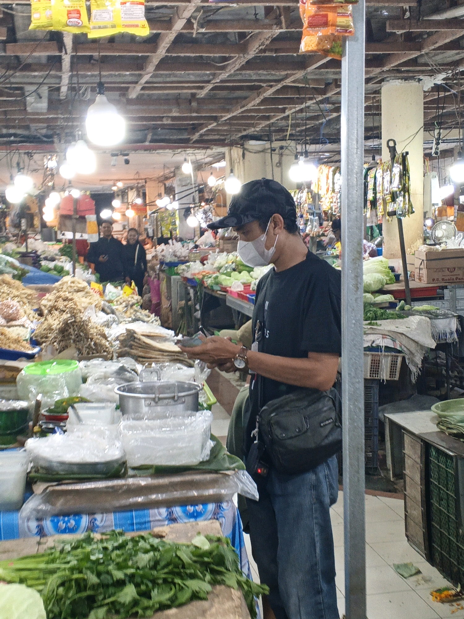 The image captures a bustling scene within a market in Denpasar, Indonesia. A man wearing a black t-shirt, blue jeans, a black cap, and a white face mask stands centrally, looking down at his phone. He has a black shoulder bag slung across his chest. Behind him, the market stalls are filled with various goods, including what appears to be dried fish, vegetables, and packaged food items hanging from the ceiling. In the background, two individuals, a man and a woman, are visible, smiling. The market is illuminated by numerous light bulbs hanging from the ceiling, casting a bright and somewhat diffuse light. The overall environment suggests a busy and active marketplace.