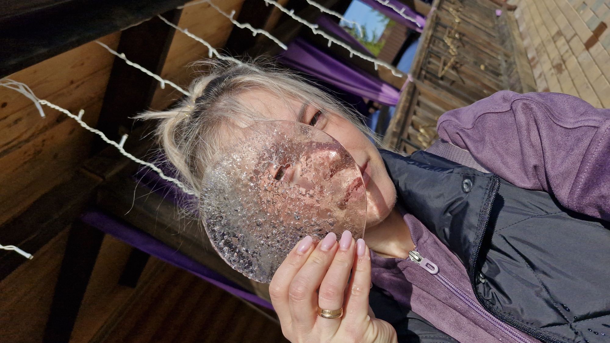 A woman with blonde hair, partially obscured by a round, clear ice formation, looks directly at the camera. The ice is held in front of her face with her right hand, which also bears a gold ring on her ring finger. She is wearing a dark, possibly black, padded jacket over a purple fleece top.
The scene is angled upwards, suggesting the photo was taken from a low viewpoint. The background consists of a partially constructed or dilapidated wooden structure, possibly a gazebo or pergola, adorned with strings of white lights. Through gaps in the wooden beams, there's a hint of blue sky and green foliage, suggesting an outdoor setting. Purple fabric is draped across the structure.
The location of Otchor, Russia, is not discernible from visual elements present in the image.