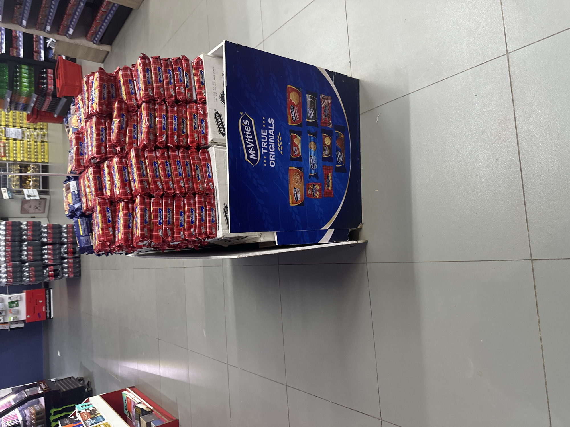 The image shows a retail environment, possibly a supermarket or convenience store, with a large display of McVitie's biscuits in the foreground. The display is a blue cardboard stand filled with stacks of individually wrapped biscuit packs, primarily featuring the "True Originals" branding. Various McVitie's product lines are depicted on the side of the display, including Hobnobs, Digestives, Rich Tea, and Cookies.
In the background, shelves stocked with bottled beverages, likely soft drinks, are visible. Other retail fixtures and aisles are partially visible, suggesting a larger store setting. The flooring is tiled in a neutral grey. The lighting appears to be typical indoor retail lighting. The image is taken from a slightly elevated, forward-facing perspective, focusing on the biscuit display. The scene depicts a commercial space in Barden Yana, Nigeria.