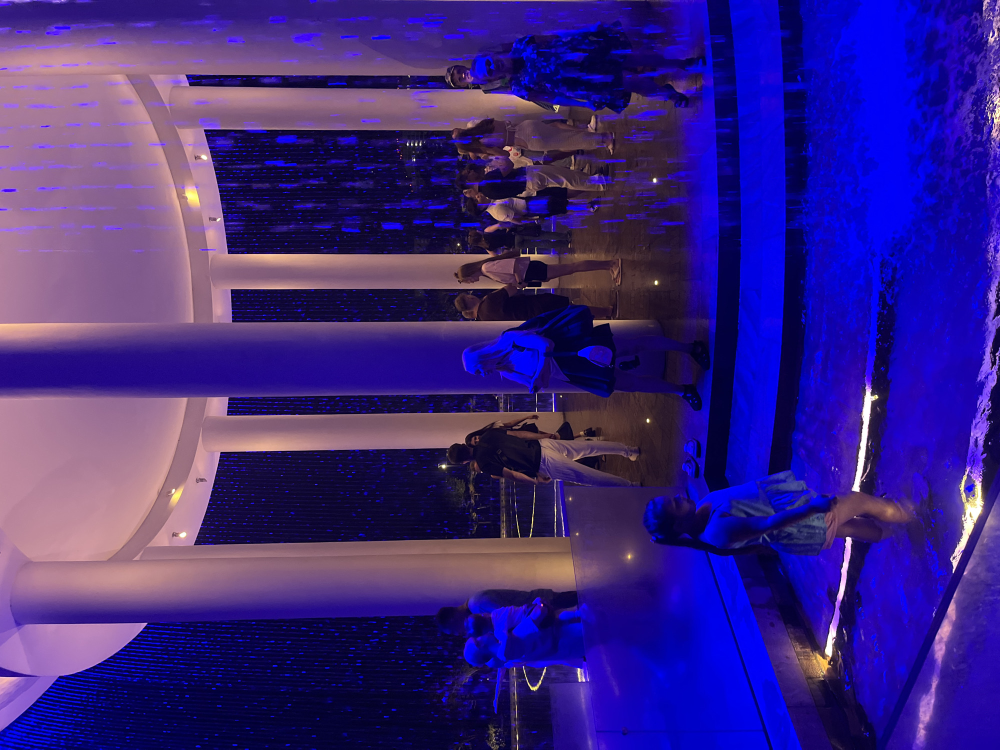 The image shows a group of people in an indoor or semi-outdoor space illuminated by blue and white lights. The scene appears to be a modern architectural setting with large white columns and a dark floor with a reflective surface. There is a water feature with cascading water visible on the right side of the image, creating a vibrant blue glow.
Several individuals are standing and walking in a line, facing away from the camera or in profile. They are dressed in casual attire, including shorts, t-shirts, and dresses. One person in the foreground on the right is partially submerged in the water feature and appears to be enjoying the experience. The overall atmosphere is calm and relaxed, possibly suggesting a leisurely outing or a visit to an attraction.
The lighting suggests it is evening or night time, and the location cues, combined with the provided context of Krasnodar, Russia, point to a contemporary public space like a park, a plaza, or an entertainment venue. There are no visible text elements in the image.