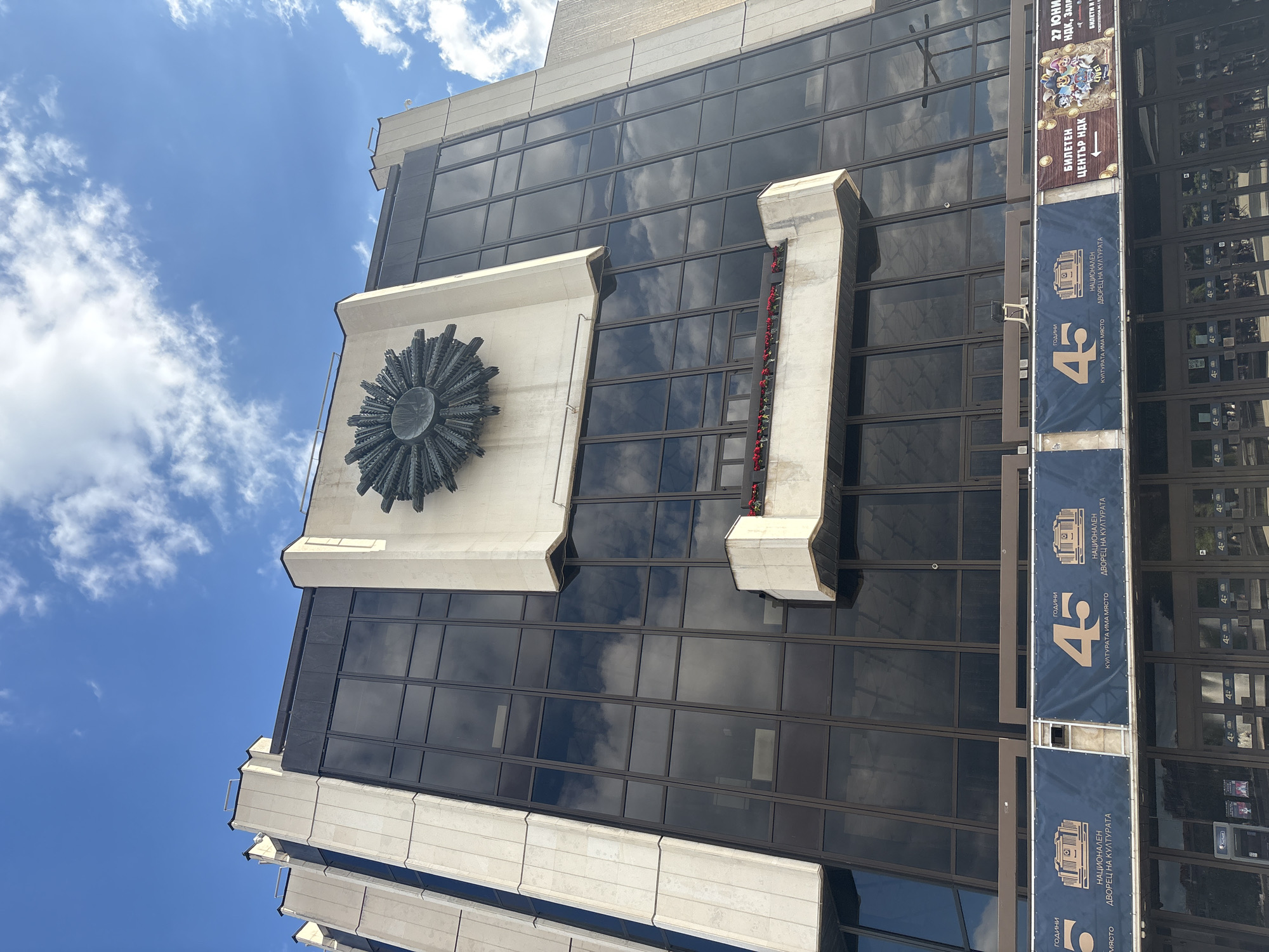 A low-angle exterior shot captures the National Palace of Culture (NDK) in Sofia, Bulgaria, under a partly cloudy sky. The Brutalist architecture of the building is prominent, characterized by its imposing concrete facade and large, dark-tinted glass windows that reflect the sky and clouds. A large, circular bronze sculpture, resembling a sunburst or a stylized flower, is mounted on a light-colored concrete panel above the main entrance. Below this, a smaller concrete ledge features a row of red flowers in planters. The building's facade is segmented by vertical concrete pillars and horizontal bands of glass. At the bottom of the image, banners are displayed across the building's lower section. One banner prominently features the number "45" with text in Bulgarian, celebrating an anniversary. Another banner advertises "Tickets" and "Live" events, with cartoon characters and event details. The entrance area shows multiple glass doors and ticketing machines. The environment is an urban setting with clear daylight and a bright sky.