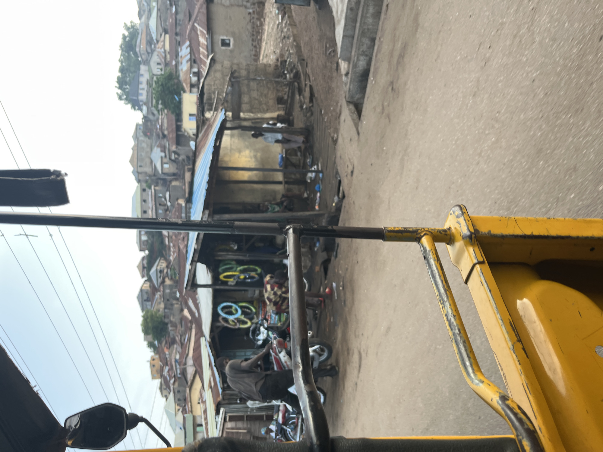 The image shows a street scene in a densely populated urban area, possibly a market or residential neighborhood, with buildings constructed on a hillside. In the foreground, a yellow vehicle, likely a tuk-tuk or auto-rickshaw, is partially visible, suggesting the photo was taken from inside or from a similar vehicle. The street is unpaved, with some scattered debris.
In the middle ground, several structures are visible, some with corrugated metal roofs, indicative of informal settlements or market stalls. A person in a gray shirt and black pants is working on a motorcycle. Nearby, several colorful tires are stacked or displayed, possibly for sale or as part of a business. Other individuals are seen near these structures, some appearing to be vendors or residents.
The background features a dense collection of houses and buildings on the hillside, with various architectural styles and roof types, including corrugated metal and what appears to be some tiled roofs. Power lines are visible crisscrossing the sky. The overall impression is one of a vibrant, active urban environment, likely in a developing country, characterized by its crowded conditions and informal economy.