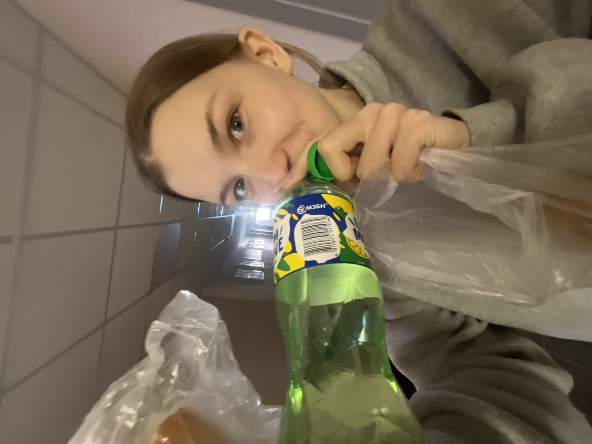 A low-angle shot depicts a woman peering into a plastic grocery bag containing a green plastic bottle of lemonade. The bottle has a yellow and blue label with the brand name partially visible as "МЗБН." The barcode on the label is identifiable as belonging to a product manufactured by the Moscow Brewing Company (МЗБН). The woman is wearing a gray sweatshirt. The setting appears to be inside an apartment building, with a glimpse of a stairwell visible in the background. The location is most likely Saint Petersburg, Russia, based on the "МЗБН" brand, which is commonly sold in Russia.