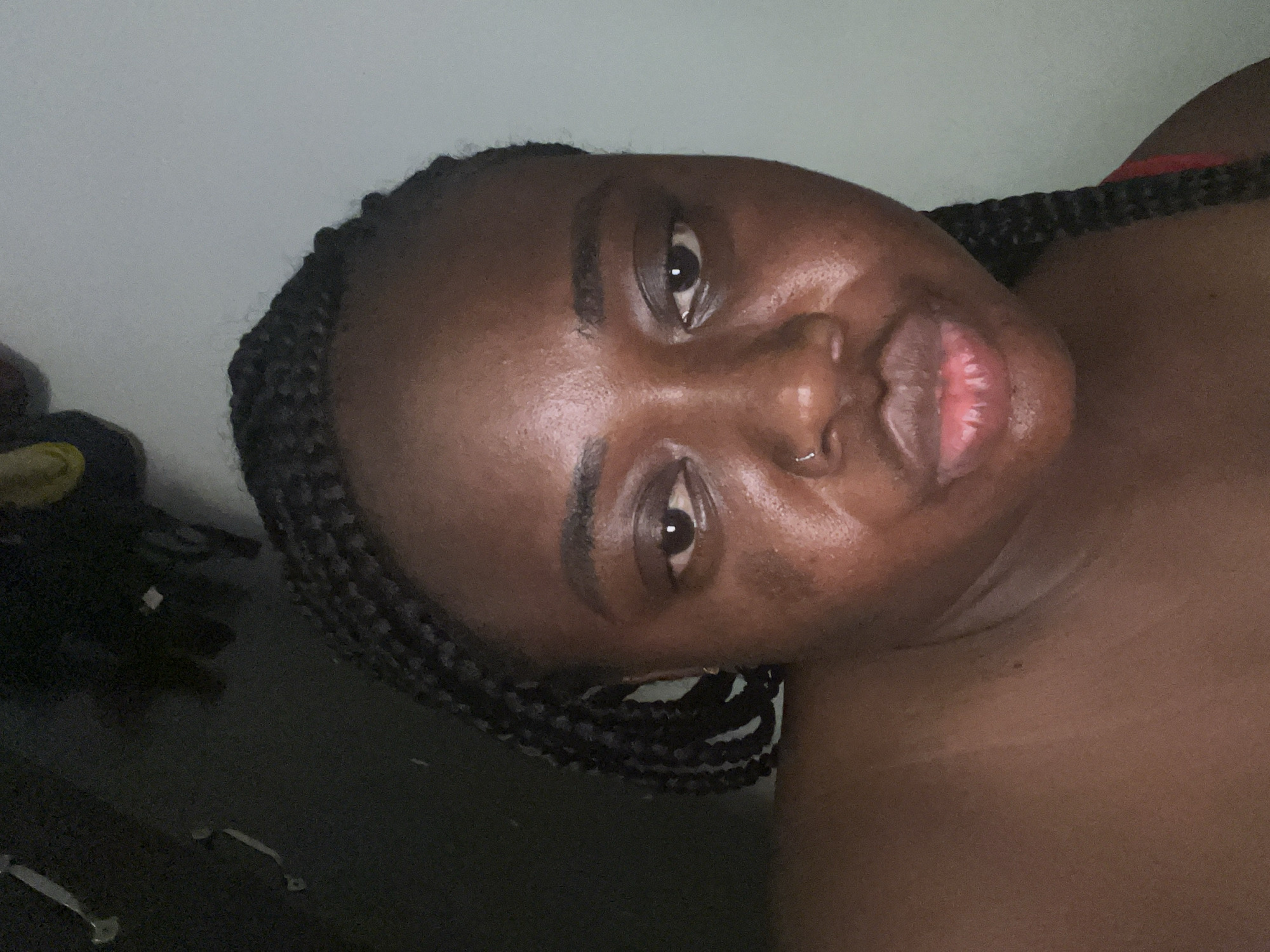 The image is a portrait of a person, likely female, with dark skin and braided hair. The subject is positioned on their back or reclining, with their head tilted to the side and eyes looking towards the camera. They have visible box braids, and a small silver nose ring is present on their left nostril. Their lips are slightly parted, and the expression is neutral or contemplative.
A red strap, possibly from a bra or top, is visible on the person's right shoulder area. The background is divided; the upper left features a plain, light-colored wall or surface, while the lower right is dark and appears to be a shadowy, indistinct area, possibly a dark piece of furniture or the floor. Faint metallic shapes resembling handles are discernible in the darker region, suggesting an indoor setting with some furniture. The lighting is soft and appears to be indoor ambient light, creating reflections on the person's skin. There is no visible text in the image.