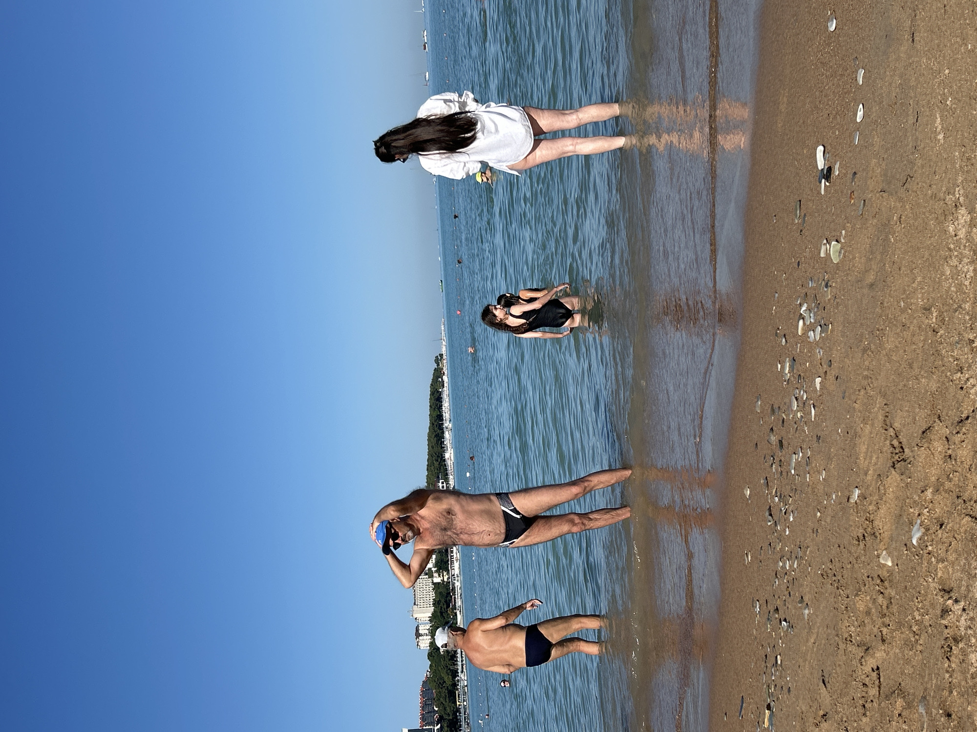 The image depicts a sunny day at a beach, likely in Gelendzhik, Russia, given the buildings visible in the background that resemble resort hotels. The water is calm and shallow, with a sandy shoreline in the foreground. Several people are present in the water.
On the left, an older man in a blue swim cap and black swim trunks stands with his back to the camera. To his right, a man wearing a blue baseball cap, sunglasses, and black speedo-style swim trunks is adjusting his cap with his hands over his head. He appears to be looking towards the right.
Further into the water, a woman with long dark hair is wearing a white, oversized shirt over a swimsuit. She is facing away from the camera and looking out towards the sea. Near her, two young girls are standing in the water. The girl closer to the foreground is wearing a black swimsuit, and the girl behind her is also in a black swimsuit.
In the distance, other people can be seen in the water, and a line of boats is anchored offshore. The buildings in the background suggest an urban or resort setting. The overall scene is one of a typical beach day with people enjoying the water under a clear blue sky.