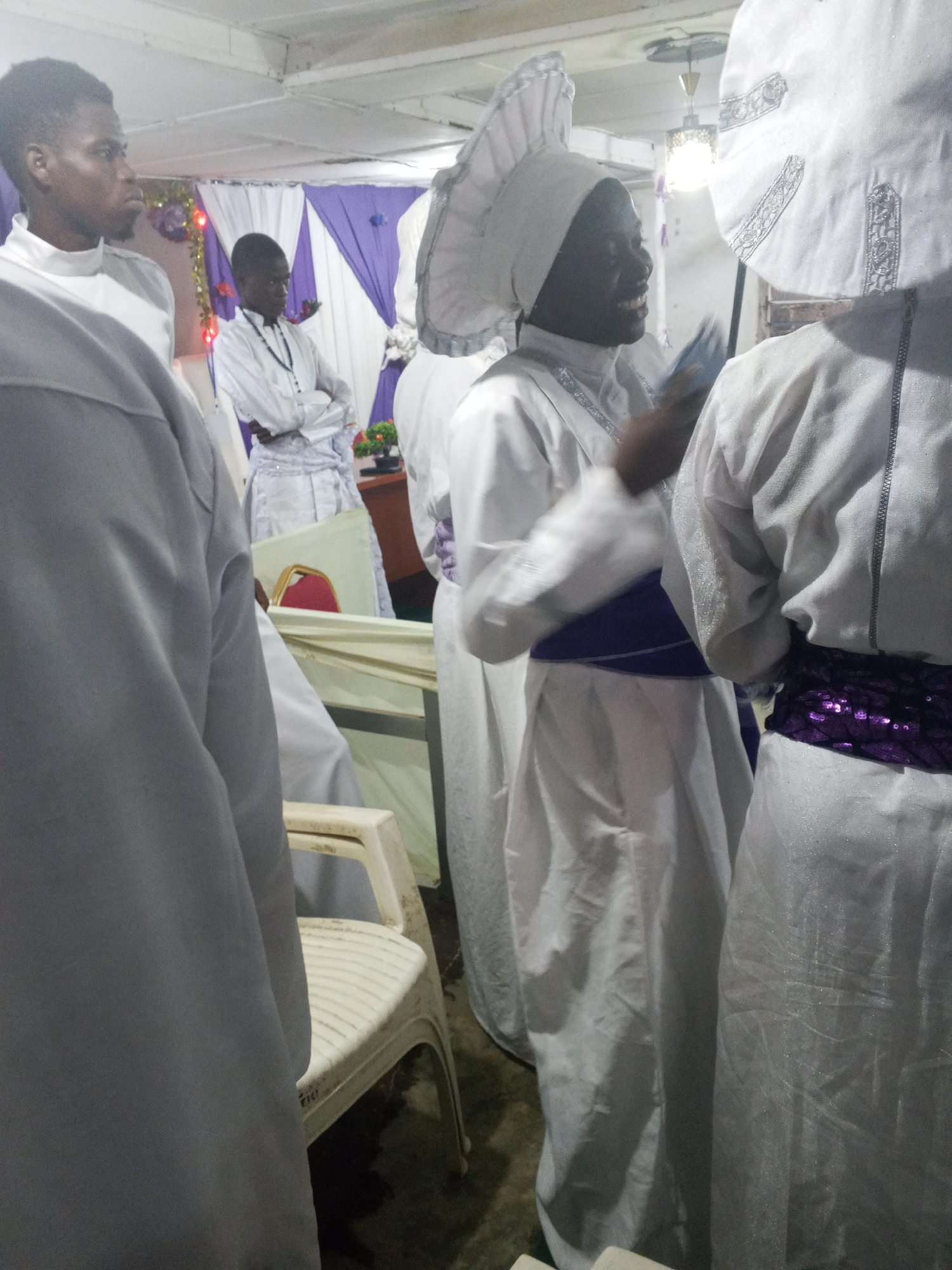 The image captures a group of individuals, likely members of a religious organization, gathered indoors. The lighting is somewhat dim, and the scene appears to be in a city identified as Unknown, Unknown. Most of the individuals are dressed in white robes and head coverings, with some adorned with purple sashes. One person is smiling and holding something in their hand. Another person stands with their arms crossed in the background. A plastic chair is visible in the foreground. Purple and white decorations adorn the background.
