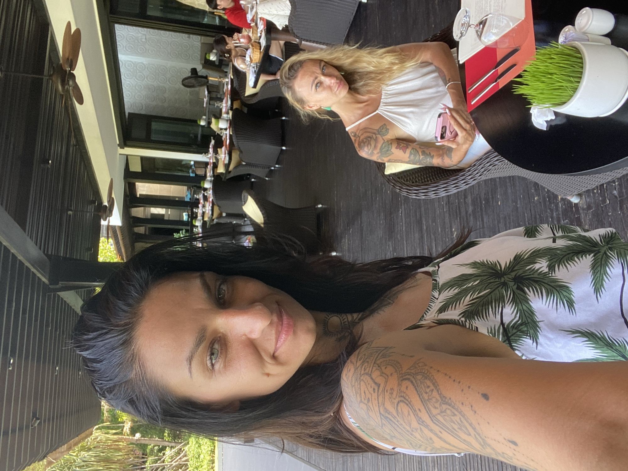 Two women are seated at tables in an open-air restaurant with dark wooden floors and a dark ceiling with exposed beams and ceiling fans. The woman in the foreground on the left is taking a selfie, her face filling the left side of the frame. She has dark hair, light green eyes, and a tattoo on her left arm and shoulder. She is wearing a white tank top with a palm tree print. The woman in the background on the right is looking towards the camera. She has blonde hair, blue eyes, and tattoos on her arms. She is wearing a white halter top. Both women have place settings in front of them on their respective tables. The woman on the right is holding a pink smartphone. The restaurant appears to be in a tropical location, with palm trees visible in the background through open doorways. The tables are set with plates, glasses, and silverware. A small pot of grass sits on the table in front of the woman on the right. The overall atmosphere is casual and relaxed. The image was taken in Phu Thuy Ward, Vietnam.