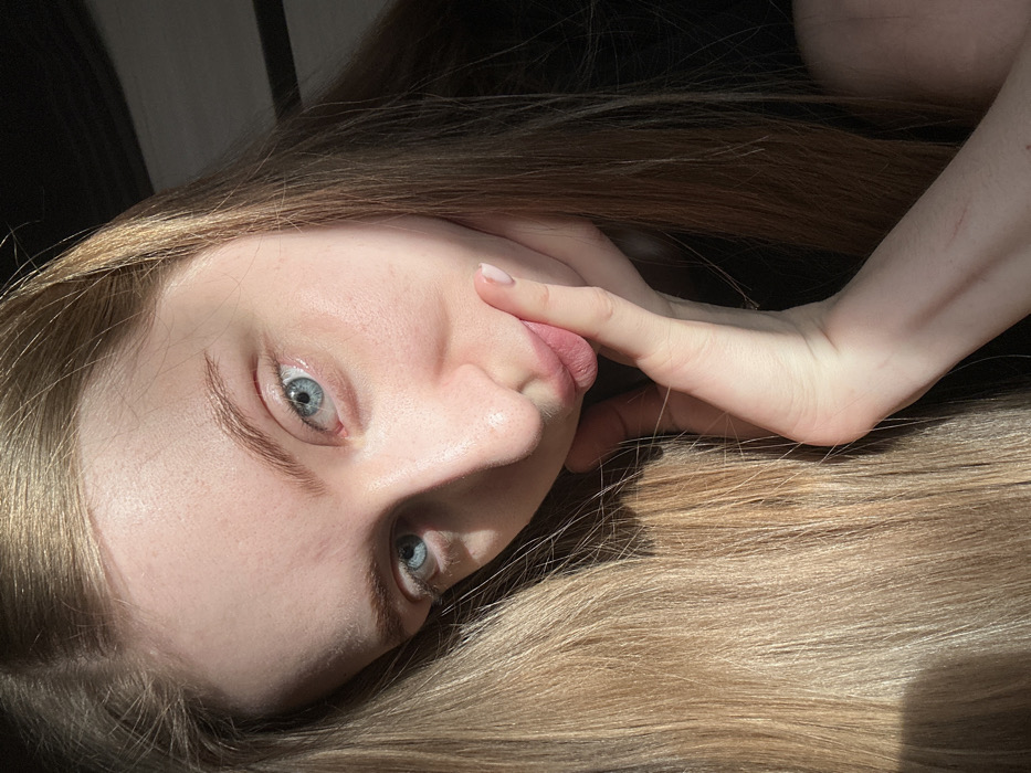 A young woman with light skin and blonde hair lies on her side, her head tilted back. Her blue eyes are prominent, and one finger touches her lips. Sunlight illuminates part of her face and hair, while shadows add contrast. The background is dark, possibly indicating a bed or a shaded area. Her arm rests on her hair, adding to the relaxed, intimate feel of the image.