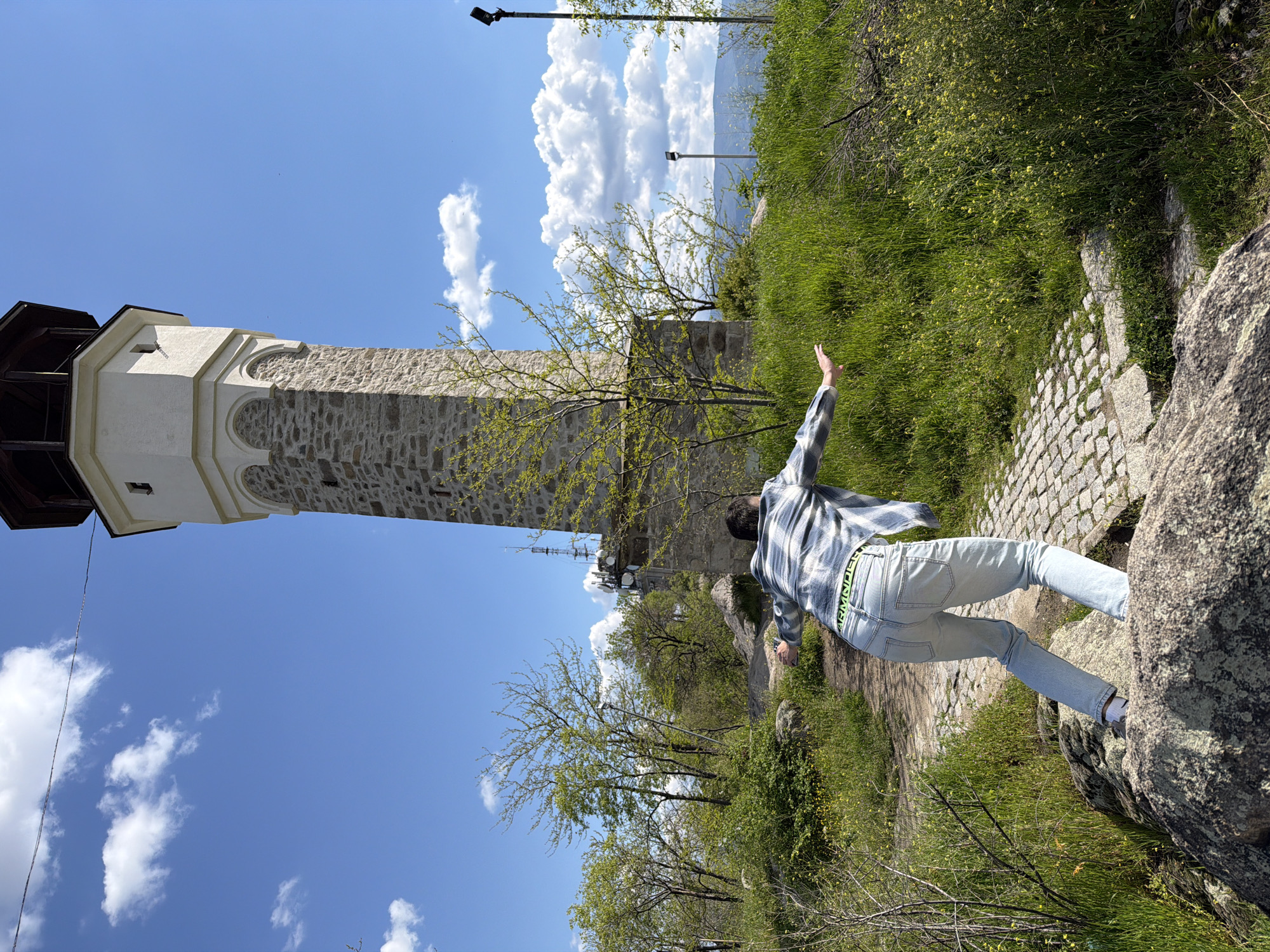 A low-angle shot features a male individual wearing a blue and white striped shirt and light blue jeans, standing on a rock formation beside a stone-paved path, with his right arm raised, captured in Unknown, Unknown. The background includes a tall stone tower adjacent to a building, along with several trees and green vegetation on a hill. The sky is blue with scattered white clouds.