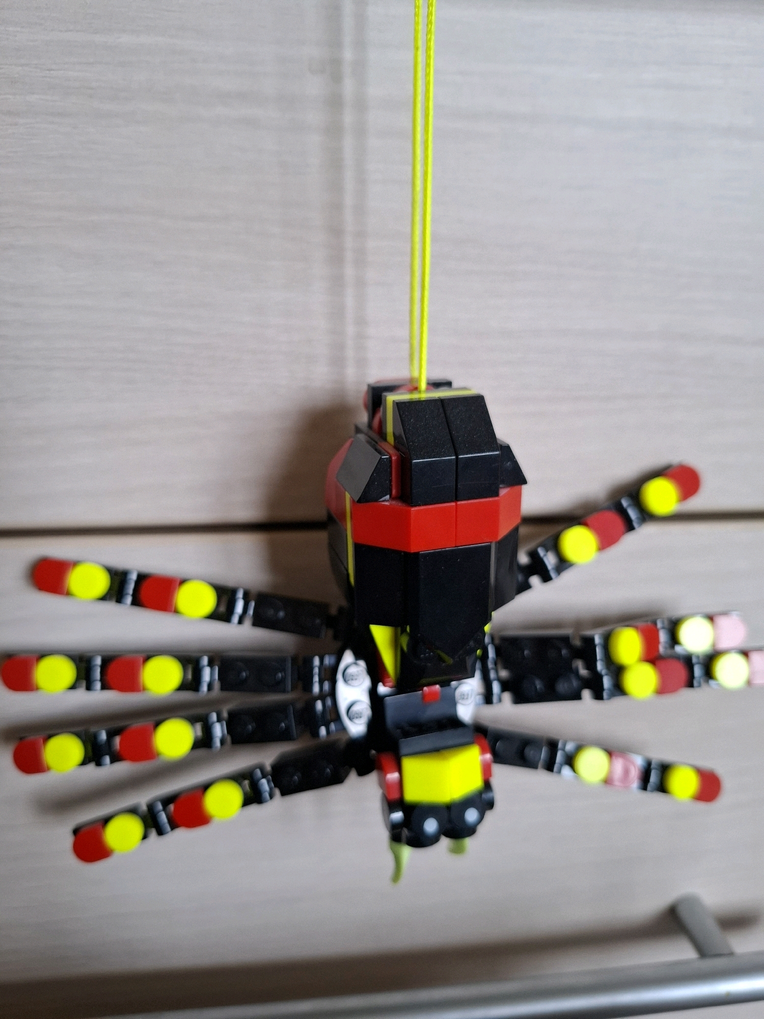 The image shows a close-up, eye-level, indoor shot of a black, red, and yellow LEGO spider hanging from a bright yellow string against a light-colored, wood-paneled background. The spider has eight segmented legs, each adorned with alternating red and yellow circular LEGO pieces. The body of the spider is constructed from black and red rectangular LEGO bricks, with a yellow section at the bottom. The background appears to be a cabinet door or wall with a horizontal wood grain pattern. The image is in focus on the LEGO spider, with the background slightly blurred. There is no information to indicate the location in Tyumen, Russia.