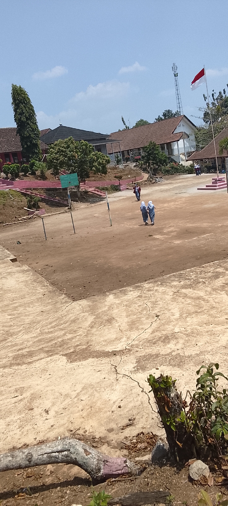 The image depicts a sunny daytime scene outside a school building, likely in Jepara, Indonesia. In the foreground, a cracked concrete surface transitions into a dusty brown dirt field, possibly a playground or sports area. A large, weathered tree trunk lies on the ground in the lower left. Two students in uniform, one in a white hijab and blue uniform and the other in a blue hijab and uniform, are walking away from the camera towards the right side of the frame. Further back, another student in a similar uniform is walking towards the left.
Behind the field, a school building with brown tiled roofs and white walls is visible. Some structures are built on a raised, terraced area with pink railings and stairs. A green basketball hoop stands on a pole in the mid-ground. In the background, a tall communication tower is visible, and an Indonesian flag with red and white stripes is flying on a pole. The sky is clear blue with a few wispy white clouds. The overall impression is one of a typical school environment in a tropical climate.