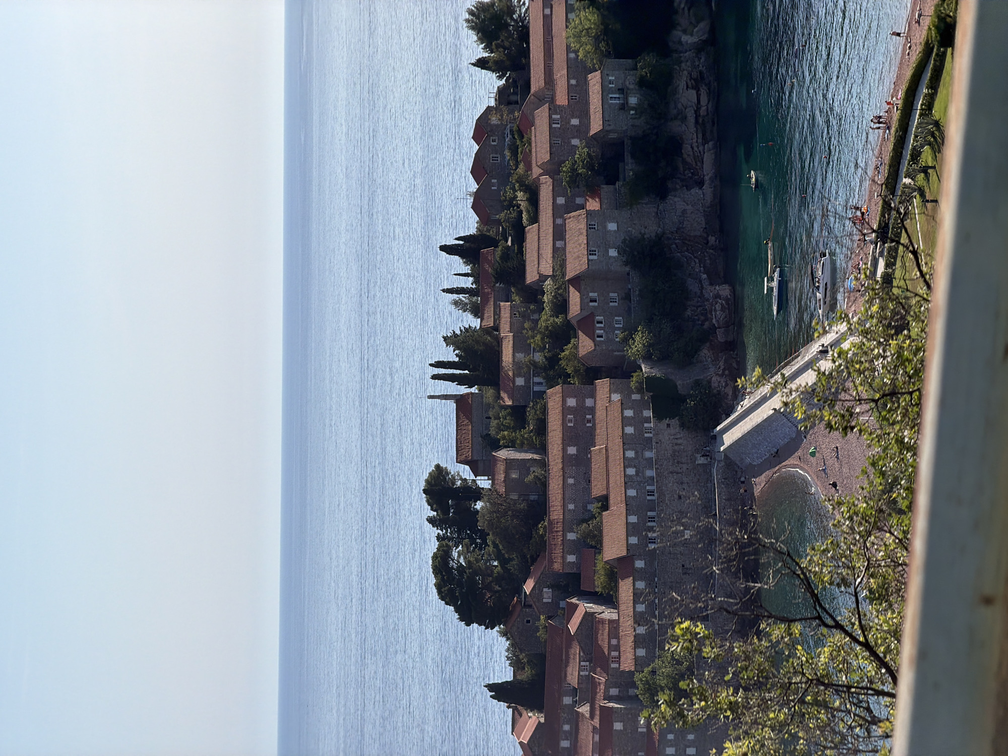 The image depicts the island city of Sveti Stefan, Montenegro, seen from an elevated vantage point. The Adriatic Sea is visible on the left side of the image. The island is densely populated with stone buildings featuring red-tiled roofs. The sea surrounds the island, and there is a small beach area visible where people are relaxing. Several boats are docked along the coastline. Trees are interspersed among the buildings. A portion of a railing and some tree branches are visible in the foreground on the right, suggesting the perspective is from a balcony or elevated observation point. The sky is clear, indicating daylight.