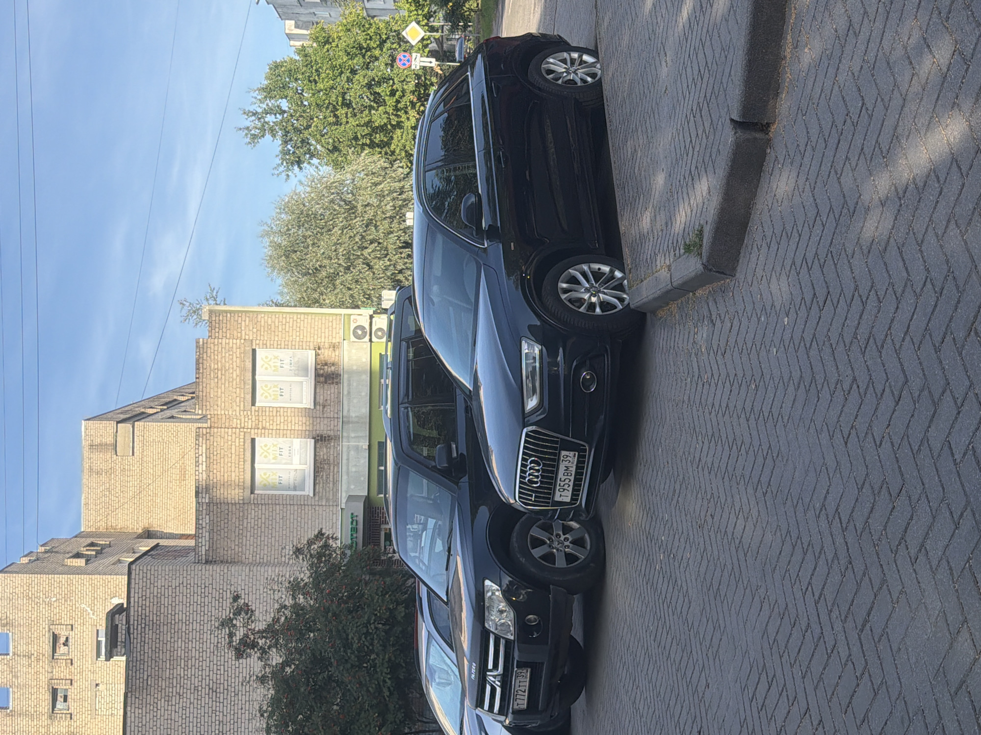 The image depicts a street scene in Kaliningrad, Russia, on a clear day with a blue sky and some clouds. Two cars are parked on a cobblestone surface, adjacent to a curb. The car on the left is a black Mitsubishi Pajero, with a license plate that reads "T 772 TT 39". The car on the right is a black Audi, with a license plate that reads "T 955 BM 39". Behind the cars, a building with light-colored brickwork is visible. The building has several windows, some of which are boarded up. There are also some trees and street signs in the background. The overall impression is of an urban environment.