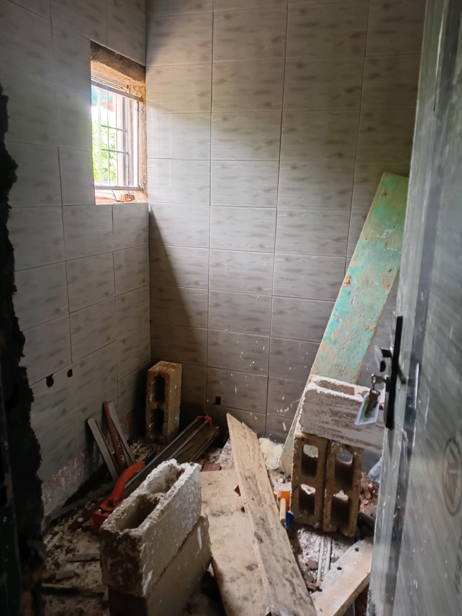 An indoor construction scene is depicted, with the walls tiled in a light grey. A small, barred window is visible on the left, allowing natural light to enter and casting shadows. The floor is covered in debris, including dirt, dust, and pieces of wood and tile. Several construction materials are present: two cinder blocks are stacked, supporting a long, green wooden plank leaning against the wall. A tile cutter with an orange handle lies on the floor near some wooden planks. In the foreground, more cinder blocks and wooden debris are scattered. A partially visible doorway with a dark door frame and a metal handle is on the right side of the image. The overall impression is that of a room undergoing renovation.