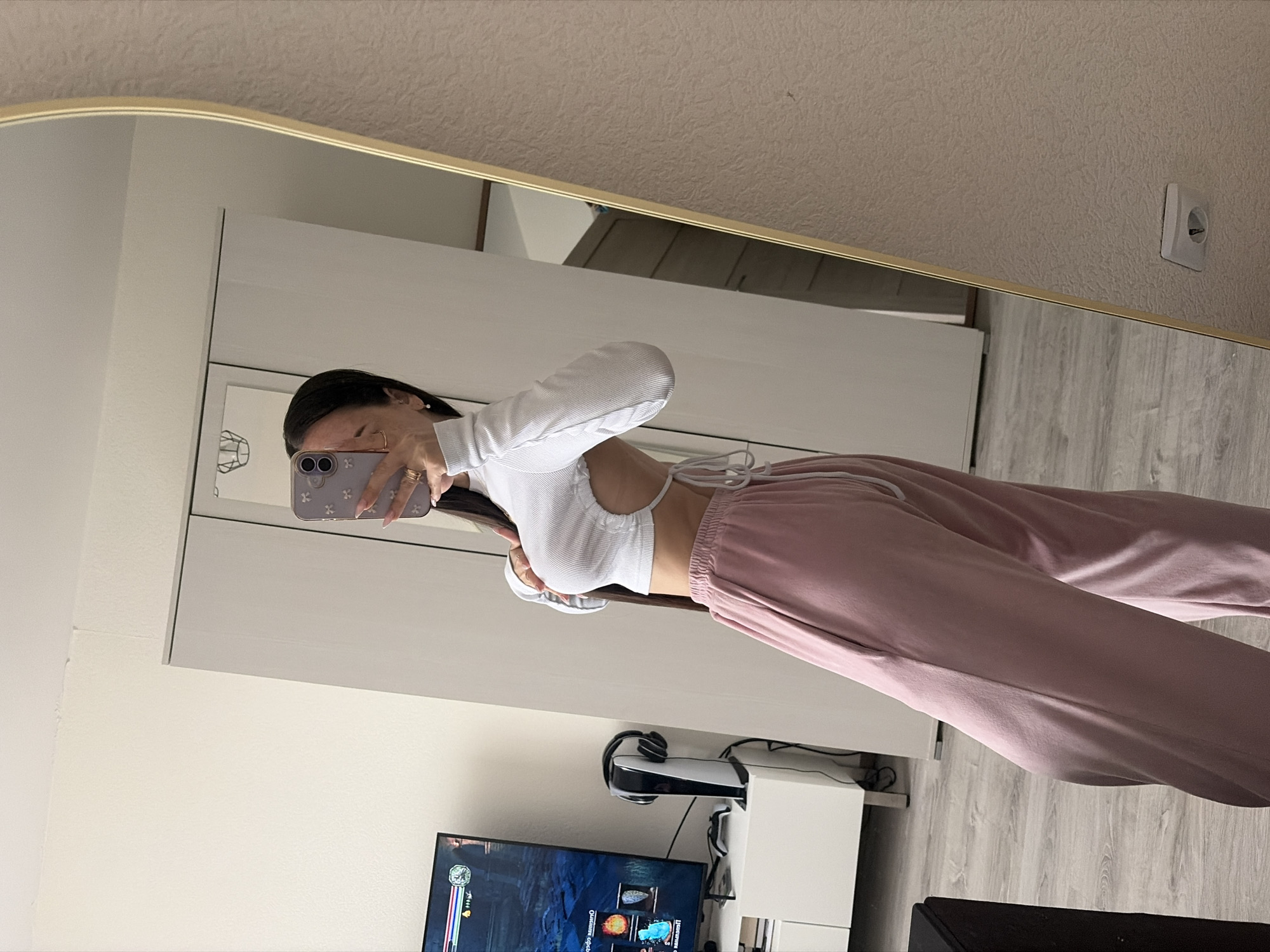 A young woman is taking a selfie in a mirror inside a room. She is wearing a long-sleeved white top with a cutout and pink sweatpants. The room contains a TV displaying a video game, a PlayStation console, a white storage unit, and wood-look laminate flooring. There is a wall outlet visible in the upper right corner of the mirror. The location appears to be a residential space, possibly an apartment. No information indicates the location is in Saint Petersburg, Russia.