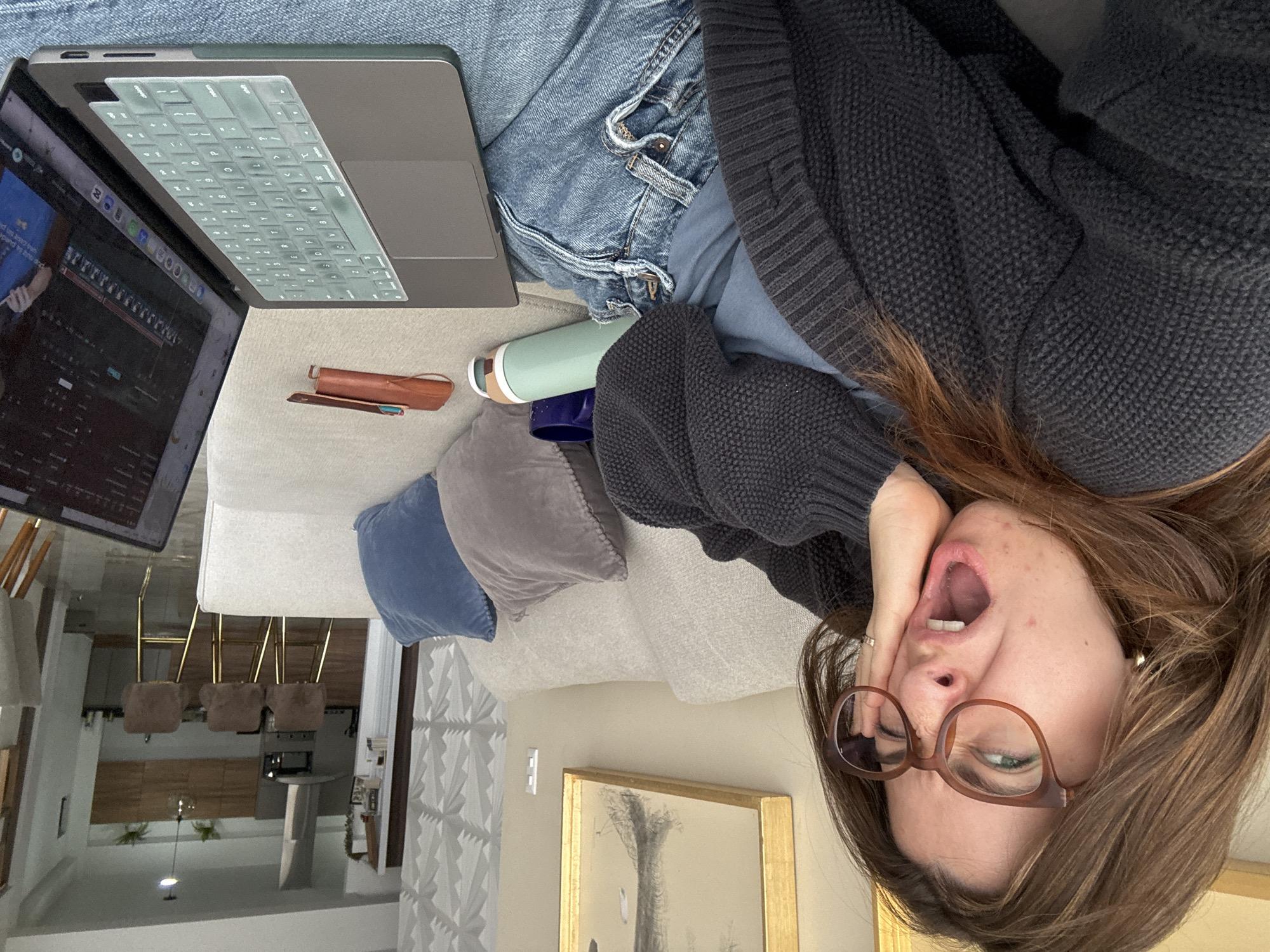 The rotated image presents a young woman lying on a light-colored couch in a modern indoor setting, likely in Puebla, Mexico. She wears blue jeans and a dark grey knit sweater over a light blue shirt. Her brown hair is spread around her head, and she wears brown-rimmed glasses. Her mouth is wide open, suggesting a yawn, while her right hand touches her cheek. A silver-colored ring is visible on her right hand.
A silver-grey laptop, likely a MacBook, rests on the couch beside her. It features a translucent, light-colored keyboard cover. The laptop screen displays a complex digital interface, appearing to be video editing software or a digital audio workstation with multiple timelines and controls. Next to the laptop, a mint green thermos or water bottle, a dark blue cup, and a small brown leather-like case (possibly a pencil case with a pen) are visible. Two decorative pillows, one light blue and one grey, are tucked into the corner of the couch.
The background reveals a contemporary living space, possibly an open-plan kitchen or dining area, with modern pendant lighting fixtures and a floor lamp. A framed artwork with a gold border hangs on a wall, and white textured wall paneling is also visible. The scene captures a moment of casual activity, possibly working or studying in a relaxed pose, with the yawn hinting at tiredness or a break in concentration. No specific legible text is discernible on the laptop screen.