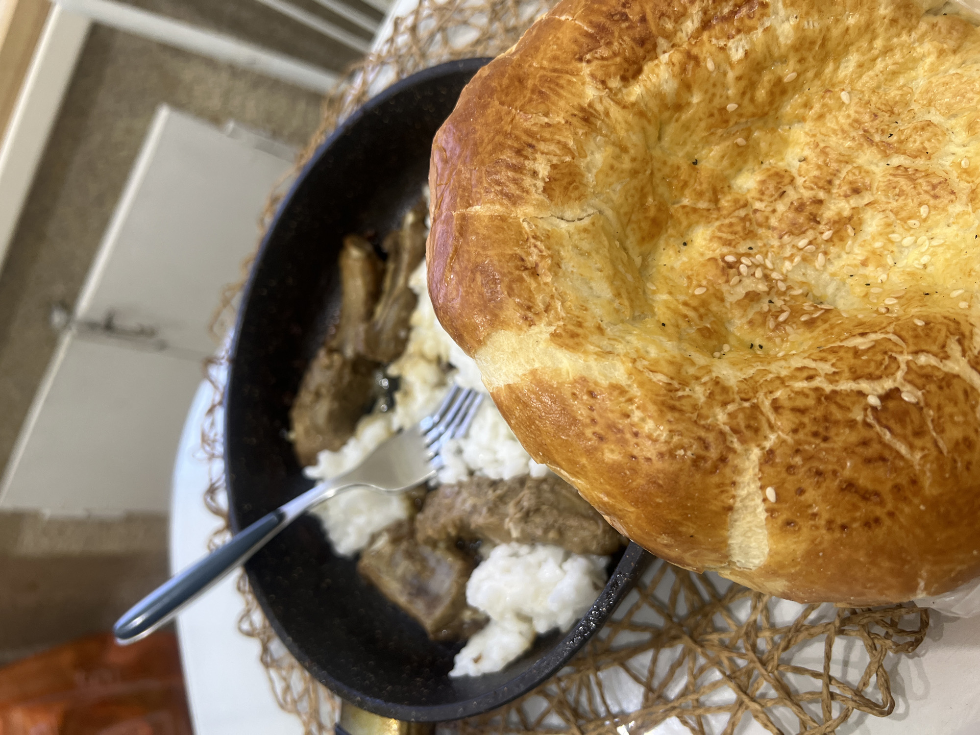 A high-angle shot shows a meal served in a black pan, with a piece of round, crusty bread to the right. The pan contains what appears to be meat and rice, served with a fork. The setting includes a woven placemat and elements of a residential or dining space. The location is not discernible from the image.