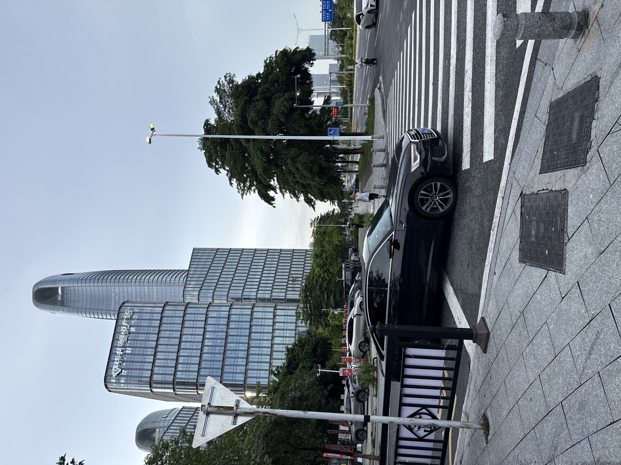 The image captures a street scene in Haizhu District, China, likely during the day. Tall, modern glass-fronted buildings dominate the background, some with curved architectural features. A large, leafy tree stands prominently in the mid-ground, casting shadows. A black car is parked on the right side of the image, partially obscuring a pedestrian crossing with white stripes.
Several people are visible: one person in a white shirt and dark shorts walks on the sidewalk near the tree, while another, wearing a white t-shirt, stands closer to the camera. In the distance, a person on a scooter or motorcycle is crossing the street.
A lamppost with two lights is visible against the light sky. The sidewalk is paved with gray tiles, and there are manhole covers embedded in it. The street is asphalt. A triangular road sign is mounted on a pole in the foreground. Several parked cars, including a white one, are lined up along the street. The overall mood appears calm and urban. Visible text on one of the buildings appears to be Chinese characters, and there are also signs with Chinese text along the street. The sky is overcast, suggesting a cloudy day.