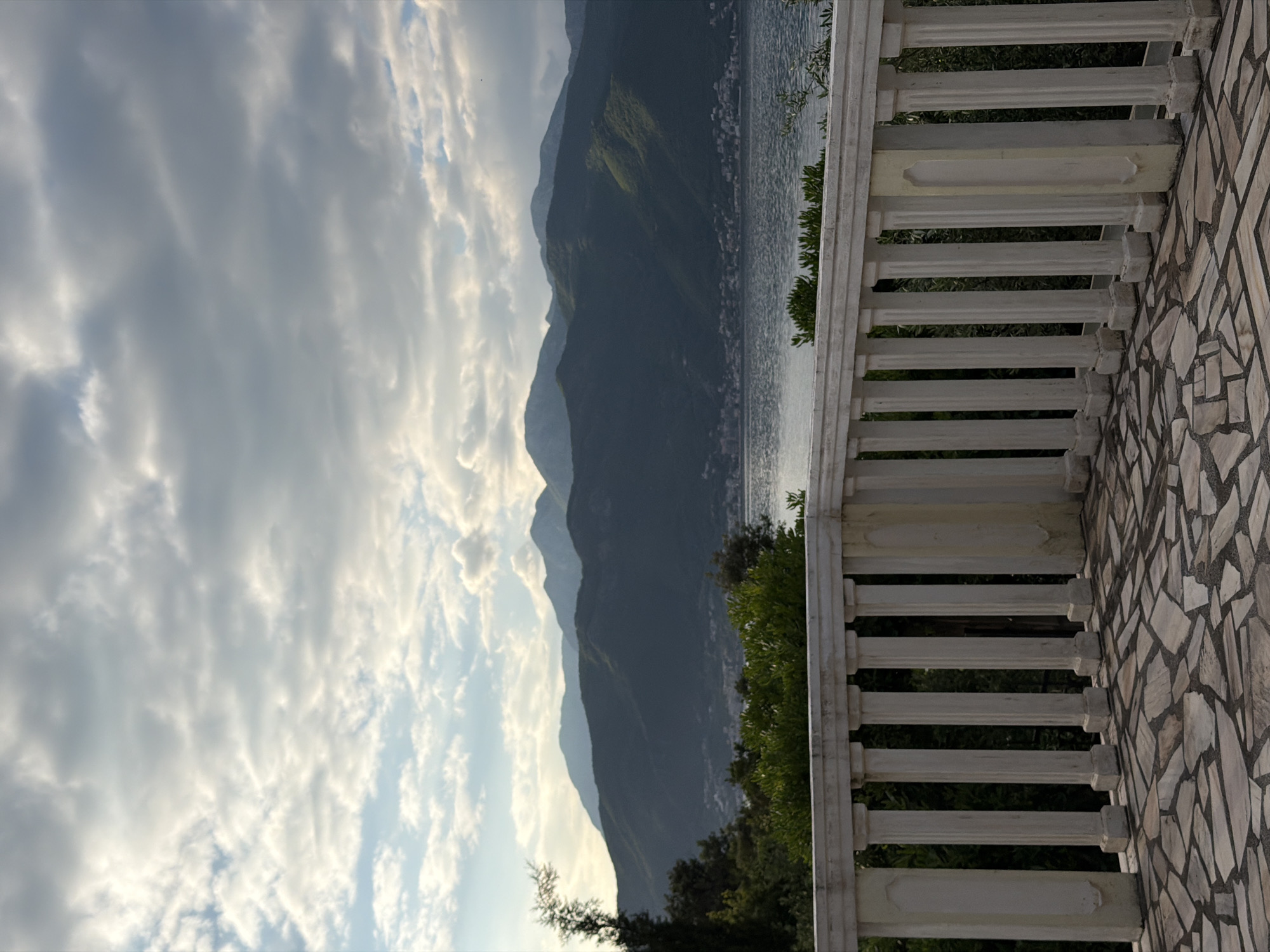 The image depicts a view from a balcony in Krašići, Montenegro. The balcony has a white, columned railing and a stone tiled floor. Beyond the railing, there is vegetation, water, and mountains. The sky is cloudy.