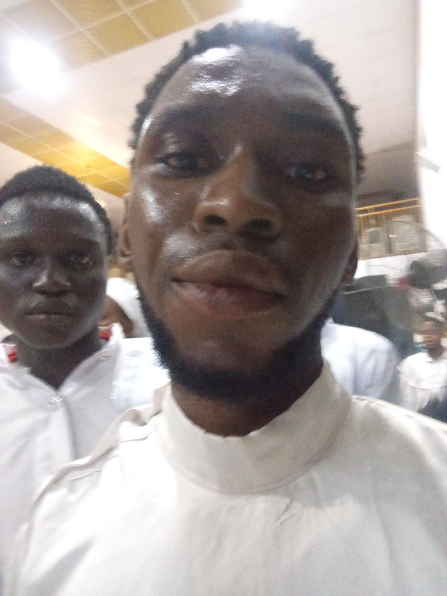 Two dark-skinned men are in a white room with a tiled ceiling. The man on the left is in a white shirt with a buttoned collar, and the other man is slightly behind him. Other people are visible in the background, also wearing white shirts. A fan is visible in the background.