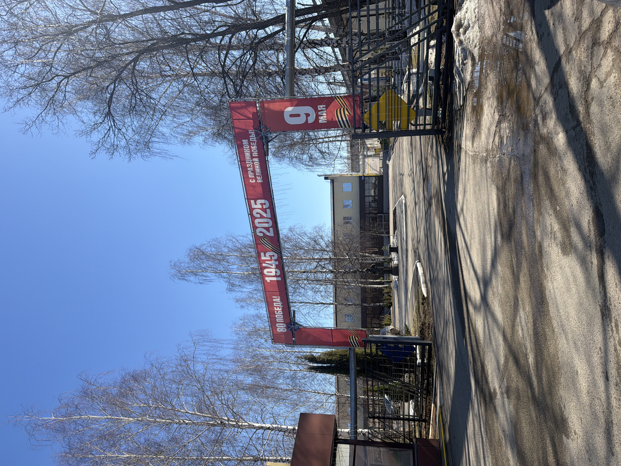 The image shows an entrance in the city of Chaikovsky, Russia. A red banner spans the entrance, displaying "9 MAY" (Victory Day) above "С ПРАЗДНИКОМ ВЕЛИКОЙ ПОБЕДЫ!" (Happy Great Victory!), "1945/2025", and "80 ПОБЕДА!" (80 Victory!). Bare trees stand on either side of the entrance. The street is paved, and a building is visible in the background.