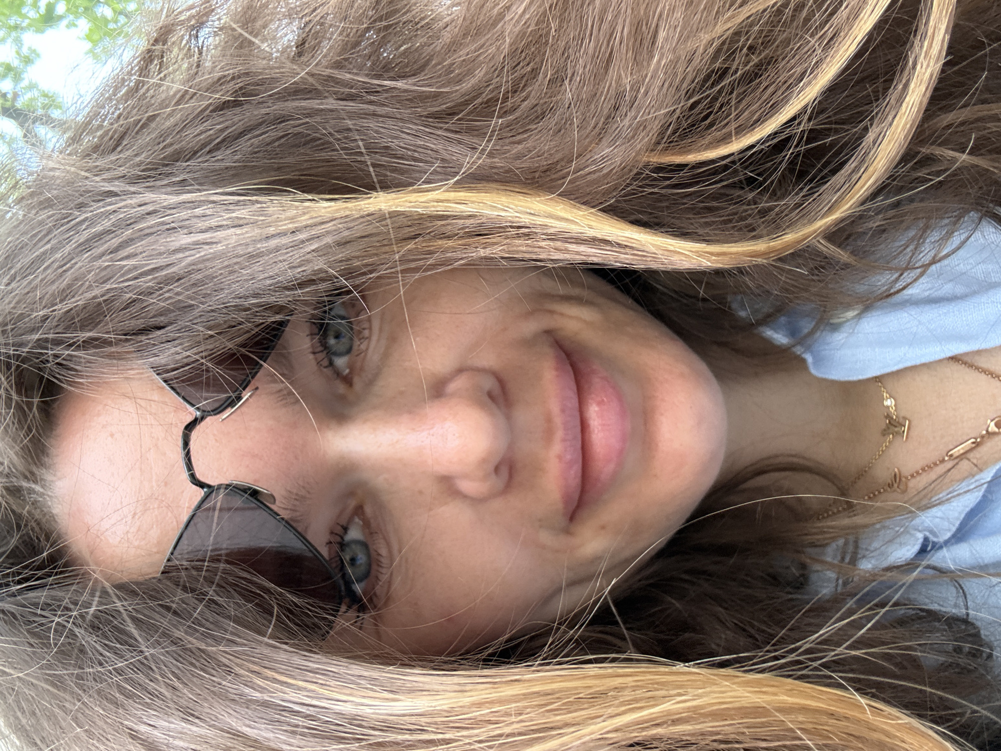 A close-up, slightly tilted portrait of a woman with long, light brown and blonde hair partially covering her face. She has fair skin, blue eyes, and is wearing sunglasses. A gold necklace is visible around her neck. The background appears to be a light blue fabric. The image does not provide any information about the location in Moscow, Russia.