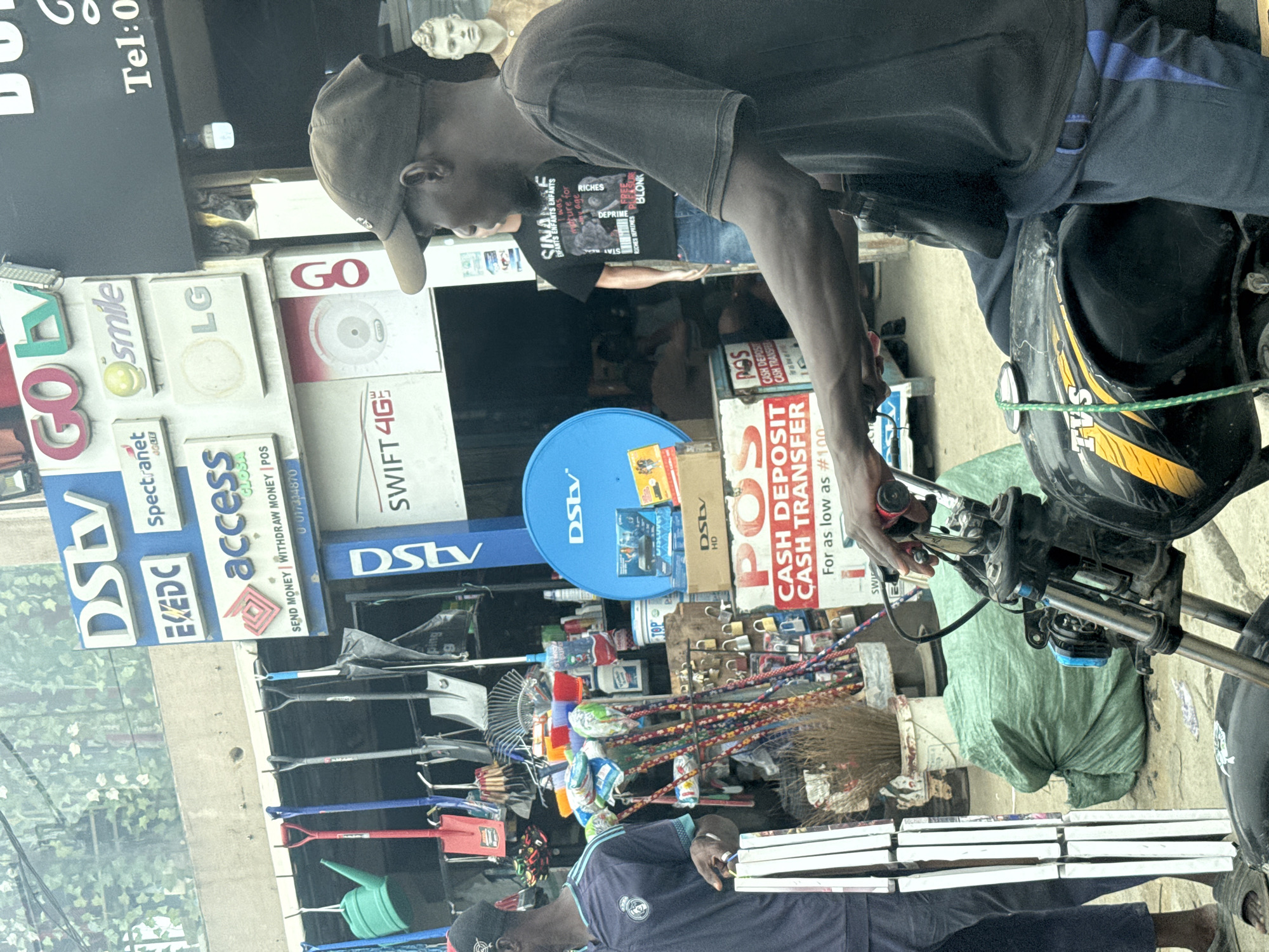 A street-level scene in Lagos, Nigeria, captured from a slightly elevated perspective, shows a bustling commercial area. The image is rotated 90 degrees clockwise.
In the foreground, a man wearing a dark t-shirt and a dark cap stands next to a black and yellow "TVS" brand motorcycle, holding its handlebars. His dark blue trousers have a light blue stripe. Partially visible behind him is a second man wearing a dark t-shirt with text that includes "SINAIS ENFANTS ENFANTS," "RICHES," "DEPRIME," and "BLONK." A third person is seen at the bottom of the frame, wearing a dark blue shirt with a white logo. On a shelf in the background, a white classical bust statue is visible.
The setting appears to be a small shop or stall, characterized by an array of signs and merchandise. Visible signs include "DSLY," "GO LY," "Spectranet," "EKEDC," "smile," "access," "LG," "SWIFT 4G," and a prominent blue "DStv" satellite dish. Other service signs indicate "SEND MONEY WITHDRAW MONEY | POS" with a phone number "0017334870," and "CASH DEPOSIT CASH TRANSFER For as low as #100." Various household and hardware goods are displayed, such as shovels, rakes, brooms, watering cans, ropes, and locks. Stacks of white rectangular objects, possibly books or frames, are seen at the bottom right.
The activity depicted is that of daily commerce and movement in a vibrant urban environment. The primary man seems to be interacting with his motorcycle, while the stall offers various telecommunication, banking, and utility services alongside physical goods. The scene occurs during the daytime, under what appears to be overcast or shaded conditions, with no specific emotions discernible from the subjects.