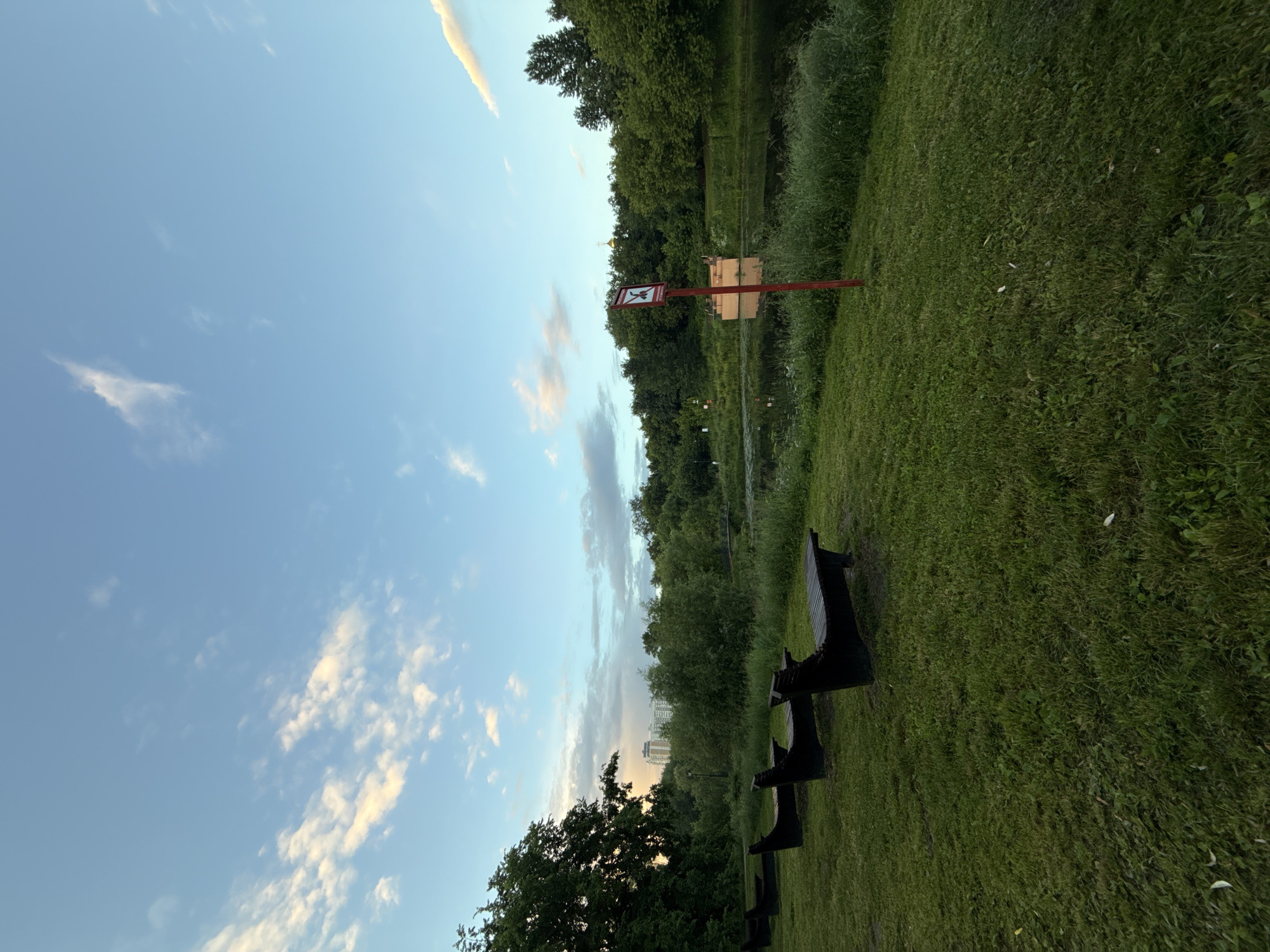 The image depicts a park scene in Moscow, Russia, during twilight. In the foreground, a lush green lawn is occupied by several dark-colored wooden benches positioned in a row, facing away from the viewer. Beyond the lawn, a body of water, likely a pond or a small lake, is visible, bordered by dense green trees and vegetation. A red pole with a sign is planted near the water's edge. The sign, a red square with a white border, features a black pictogram of a person with a line through it, indicating a prohibition. In the background, several taller buildings, characteristic of a city skyline, are faintly discernible through the trees, along with the golden dome of a church. The sky above is a soft blue, streaked with wispy white and orange clouds, suggesting the last moments of sunset. The overall atmosphere is serene and tranquil.