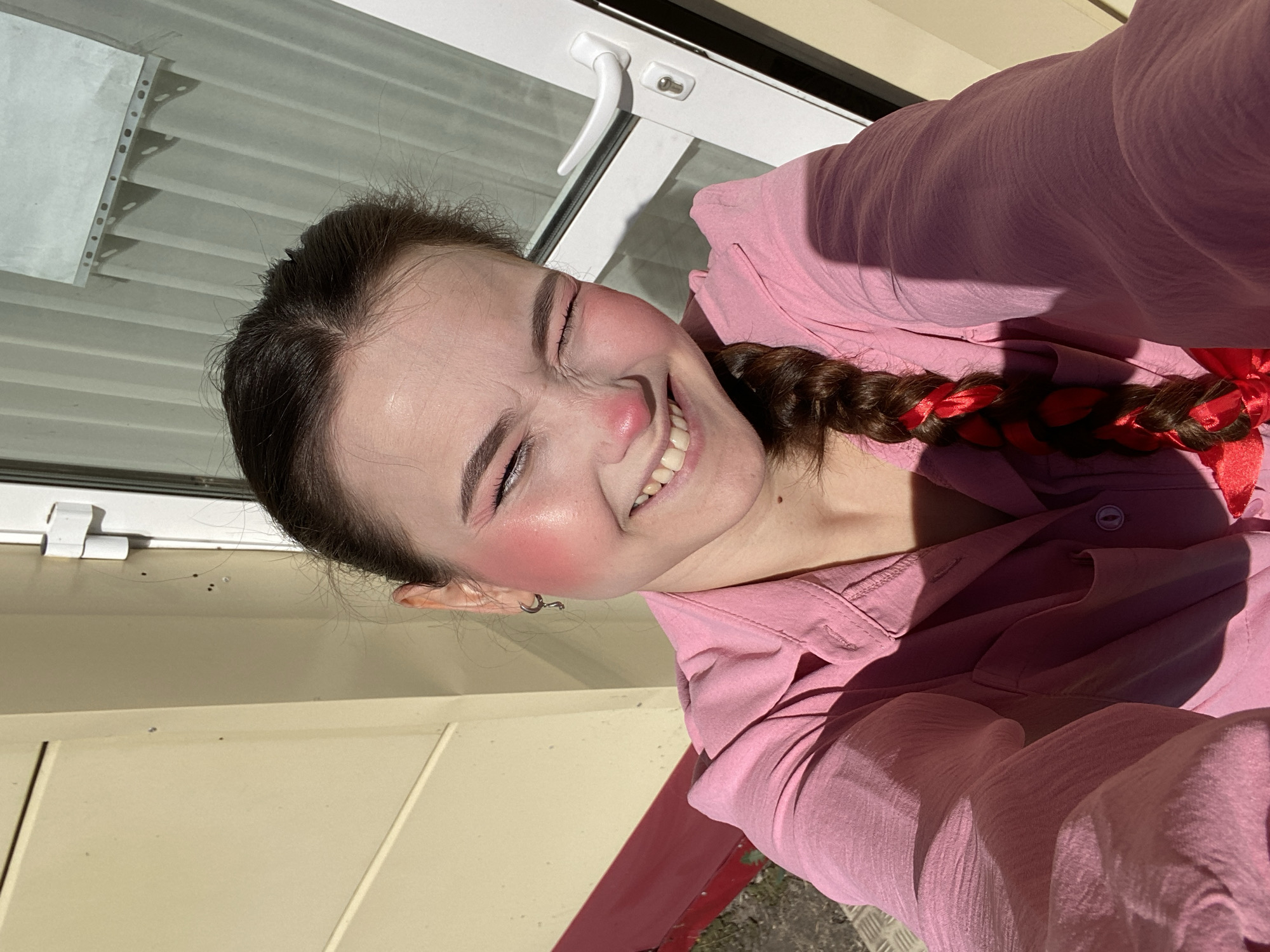 A young woman with dark brown hair in a braid adorned with red ribbons, is taking a selfie outdoors. She is wearing a pink collared shirt. She has bright pink blush on her cheeks and nose and is smiling widely with her eyes closed. The background shows a cream-colored building with horizontal siding and a white door with a glass pane and a white handle. A red railing is visible below the building. The overall lighting suggests it is daytime with strong sunlight creating shadows. The location is on a street in the city of Chaikovsky, Russia.
