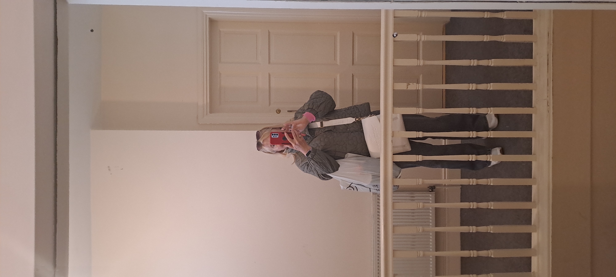 A full-body mirror selfie is captured in what appears to be an indoor hallway. The subject, a fair-skinned woman with blonde hair and sunglasses perched on her head, is holding a red smartphone to take the picture. She is wearing a quilted olive-green jacket over a pink long-sleeved shirt, dark-colored trousers, and white sneakers. A white cross-body bag with embossed lettering and a white plastic shopping bag are slung over her left shoulder. The background shows a light-colored wall, a white-paneled door, a radiator, and a wooden railing. The floor is covered in dark carpet. The environment suggests a residential or institutional setting, possibly a home or a hotel hallway. There is no indication of the specific location within Castlebar, Ireland.