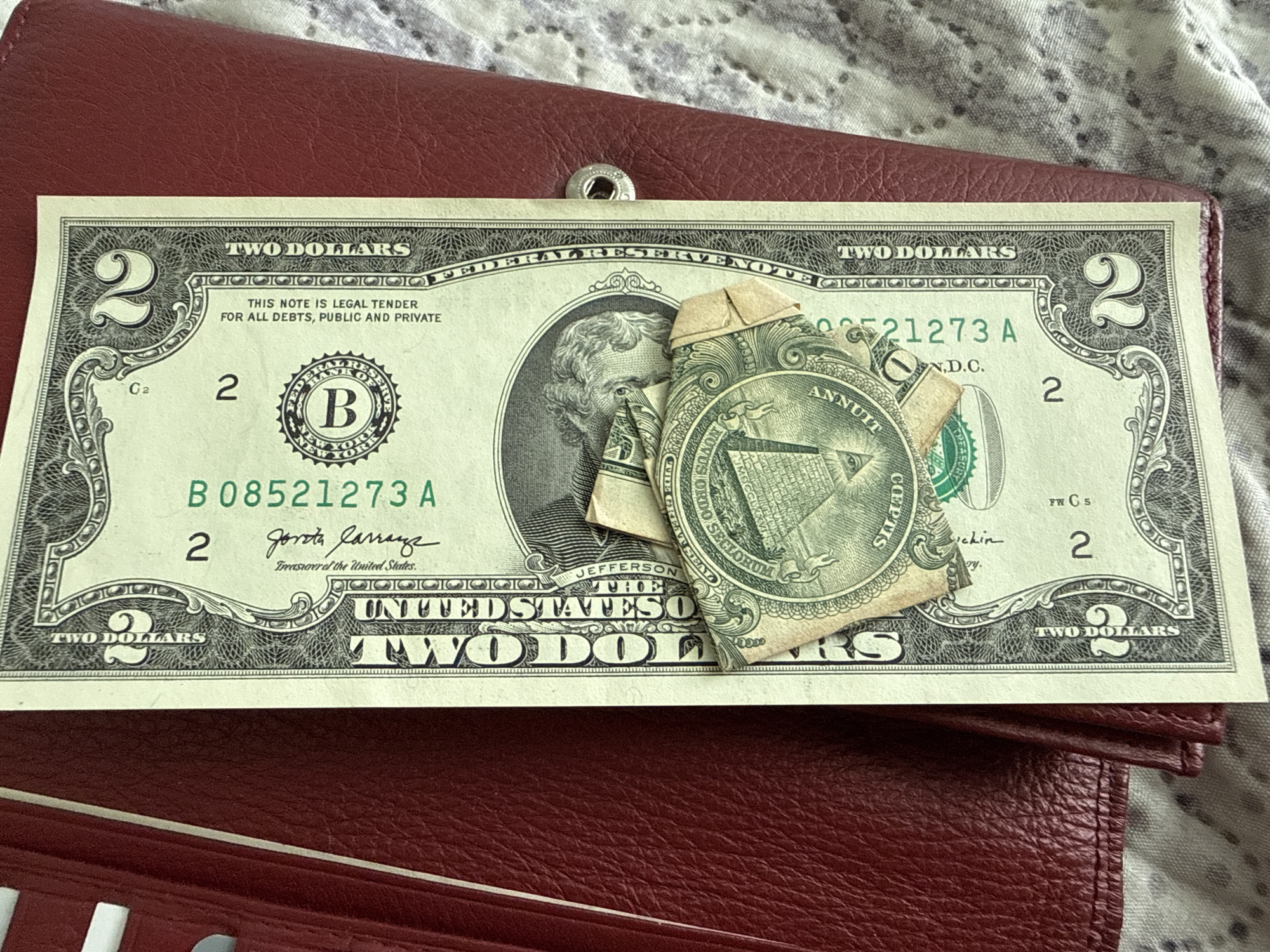 A US two-dollar bill is partially visible, lying on a red textured surface, possibly a wallet. The bill features the portrait of Thomas Jefferson and various seals and text. A portion of a one-dollar bill, folded, is on top of the two-dollar bill, showing an eye within a pyramid. The image is taken from a top-down perspective, with a slight shadow cast on the bill. There are no visible people, actions, or interactions depicted. The environment beyond the bill and wallet is out of focus, showing a patterned fabric. There is no information in the image that can determine its location in the city of Ekzarh Antimovo, Bulgaria.