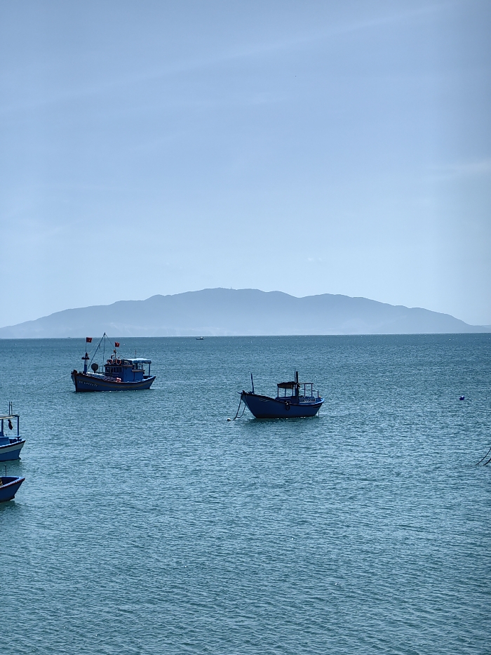 The tranquil turquoise sea hosts several blue fishing boats. A larger boat, complete with a red flag and visible equipment, is positioned to the left. A smaller boat floats near the center, gently tethered. A partially visible boat sits on the bottom left. A distant, hazy mountain range stretches across the horizon under a clear, light blue sky.