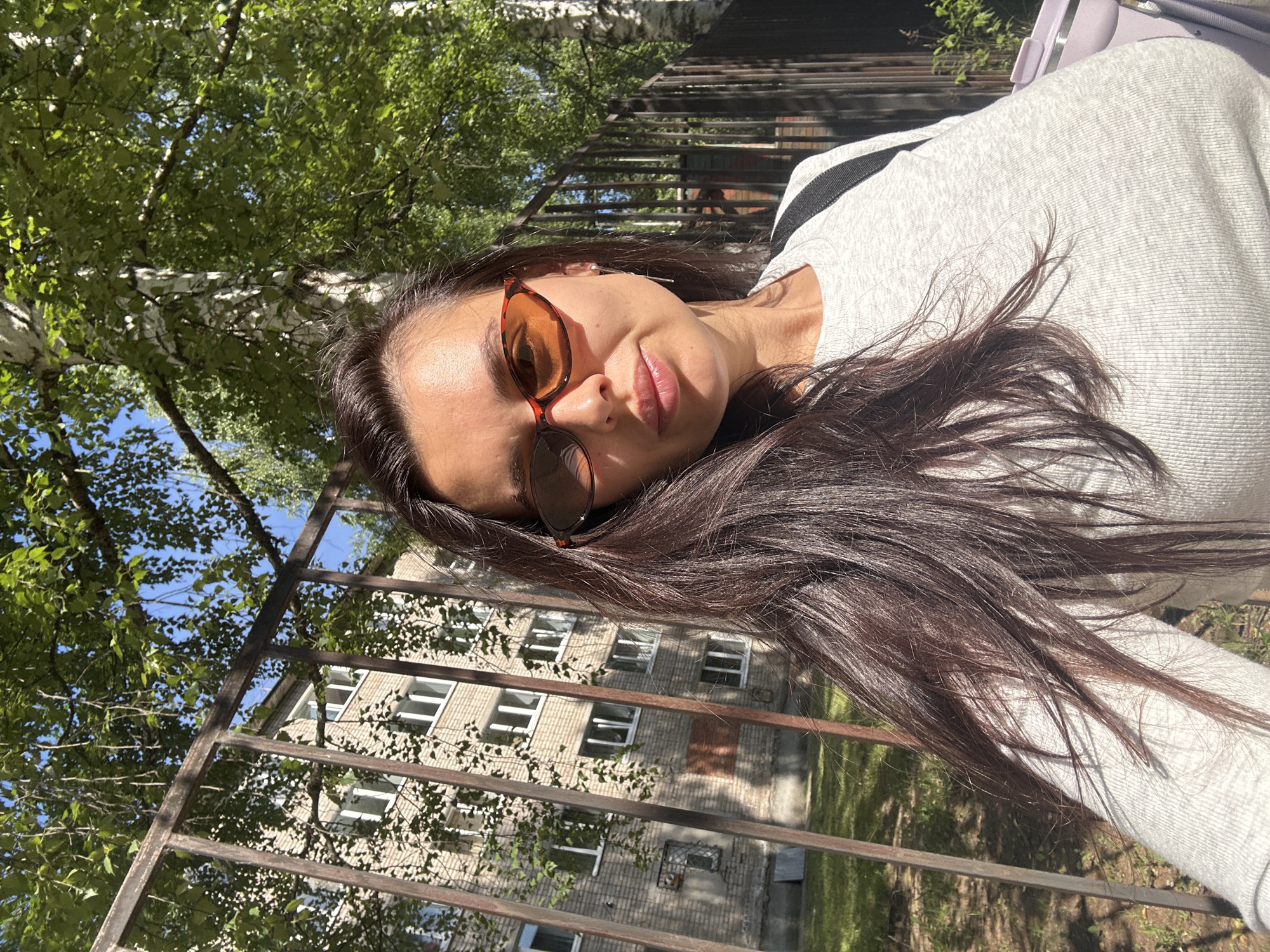 A young woman with long, dark hair and sunglasses takes a selfie outdoors. She is wearing a light gray sweatshirt with a black stripe. The sun is casting strong shadows, indicating it is a bright, sunny day, likely mid-day. She appears to be outdoors, with trees and a fence in the background. Behind the fence, a multi-story brick building with many windows is visible, suggesting an urban or suburban setting. The woman's expression is relaxed, with a slight smile. The orientation of the image suggests the photographer may have been lying down or leaning. No visible text is present in the image. The location context of Chaikovsky, Russia, places this scene within a specific geographical area.