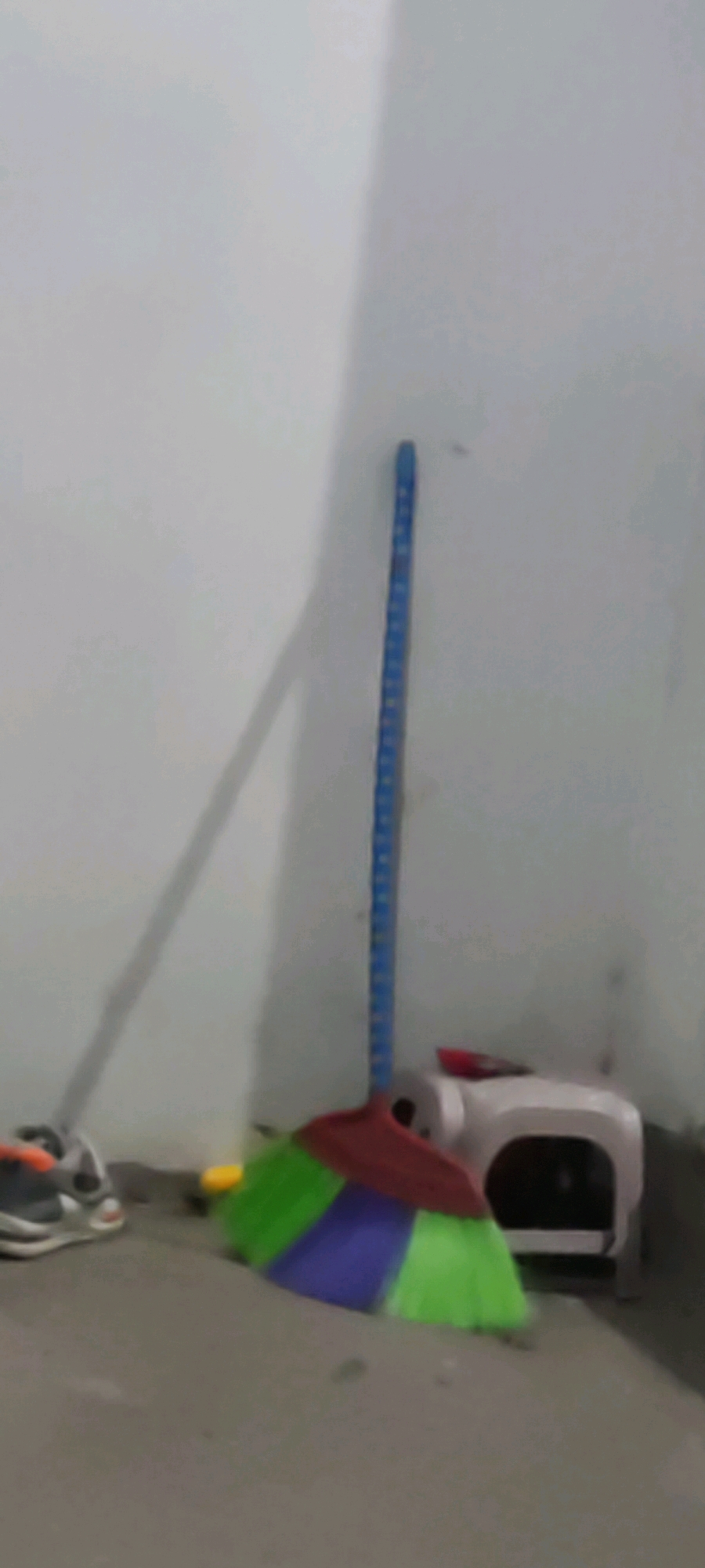A broom with a blue handle and multi-colored bristles (green, purple, and red) stands upright in the corner of a room. To the left of the broom, a pair of grey and orange athletic shoes are visible. A small, white plastic stool with a black opening is positioned behind the broom. The room has white walls and a grey floor. A shadow of the broom handle stretches across the left wall. The scene appears to be indoors, possibly in a domestic setting or a utility area, within the city of Karangwangun, Indonesia.