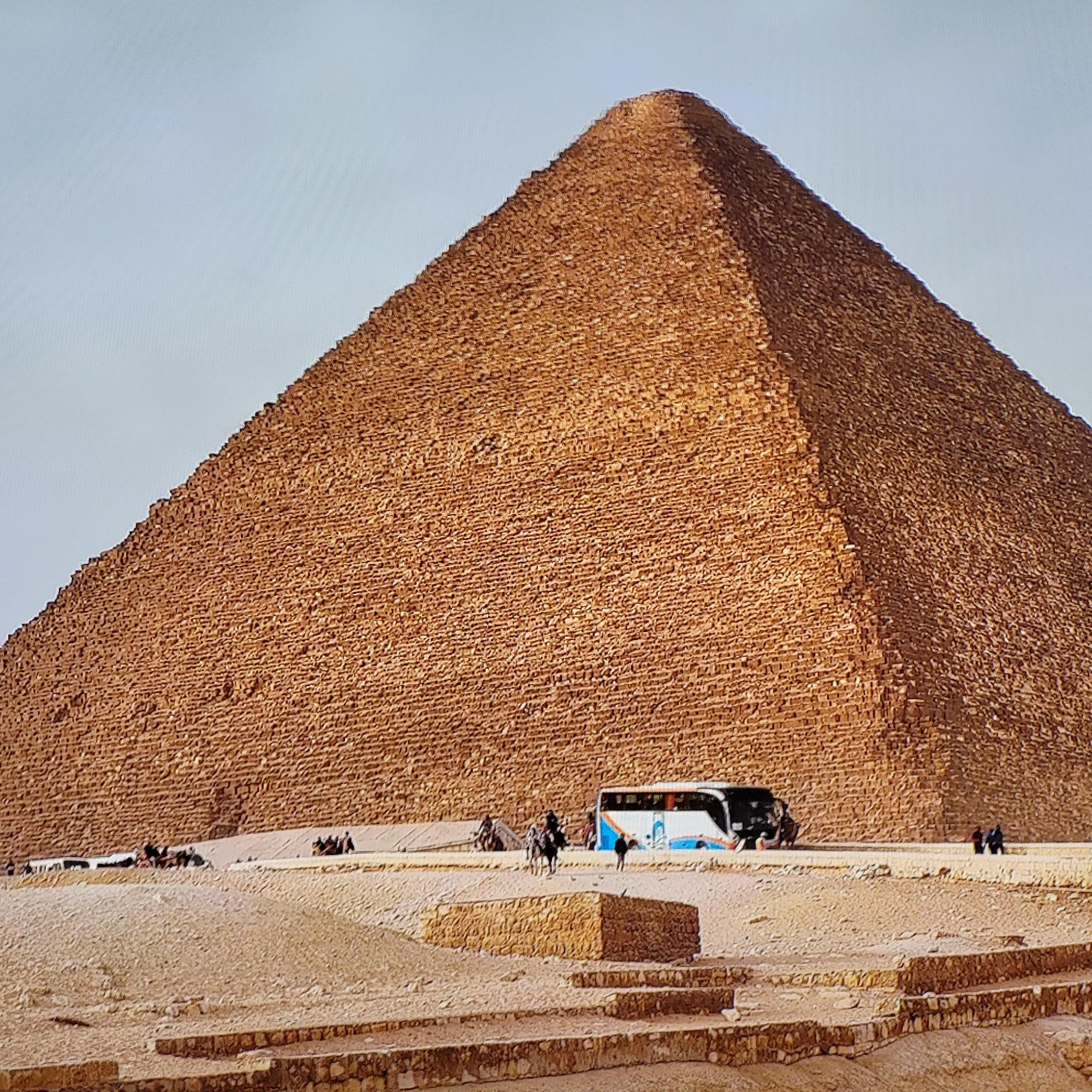 The image shows the Great Pyramid of Giza in Egypt. The pyramid is a massive structure made of stone blocks, dominating the scene. In the foreground, there is a sandy desert landscape with some low stone walls or steps. Several people are visible in the middle ground, along with a tour bus. Some people are riding horses or camels. The sky is pale and clear. The image captures the scale and setting of the pyramid.