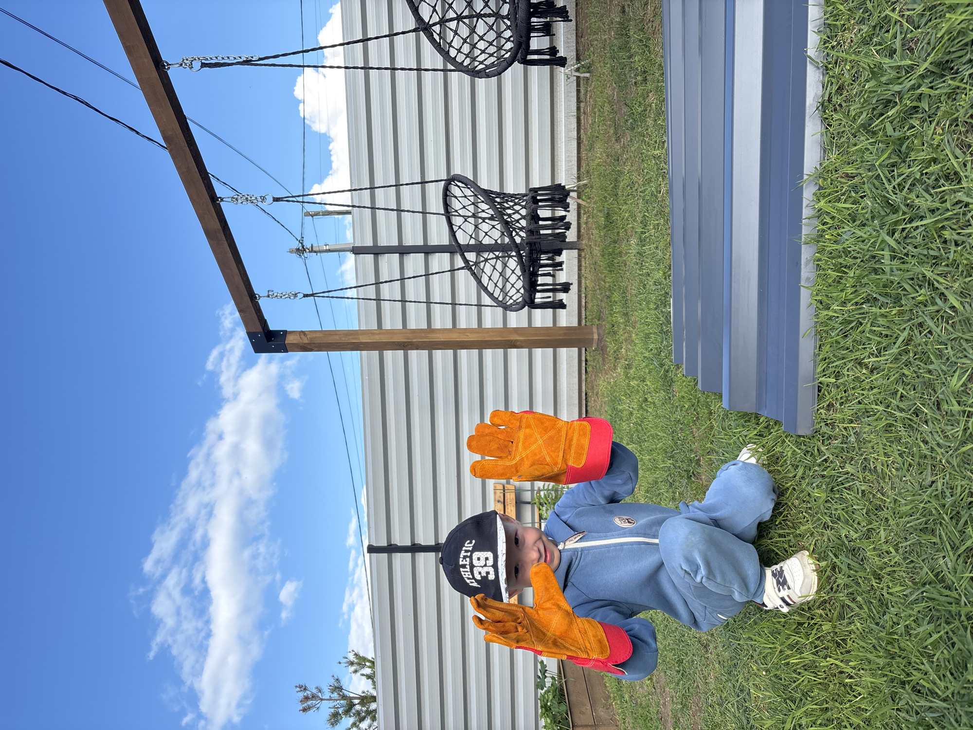 A young child is seated on vibrant green grass outdoors on a sunny day. The child, likely a toddler or preschooler, is wearing a blue jumpsuit, white sneakers, and a black baseball cap with "ATHLETIC 39" emblazoned in white text. They are adorned with oversized orange and red work gloves, which they hold up, palms facing the viewer, as they look directly at the camera with a slight smile.
In the background, a tall, light-colored corrugated metal fence runs horizontally. Above the fence, a wooden beam structure is visible, from which metal chains and black ropes suspend two black woven swing seats. A clear blue sky with scattered white clouds occupies the upper portion of the frame. To the right of the child, on the grass, lies a blue-grey corrugated panel. The scene suggests a backyard or an outdoor play area in Chaikovsky, Russia, characterized by bright daylight and good weather. The large gloves on the small child create a notable, playful detail.