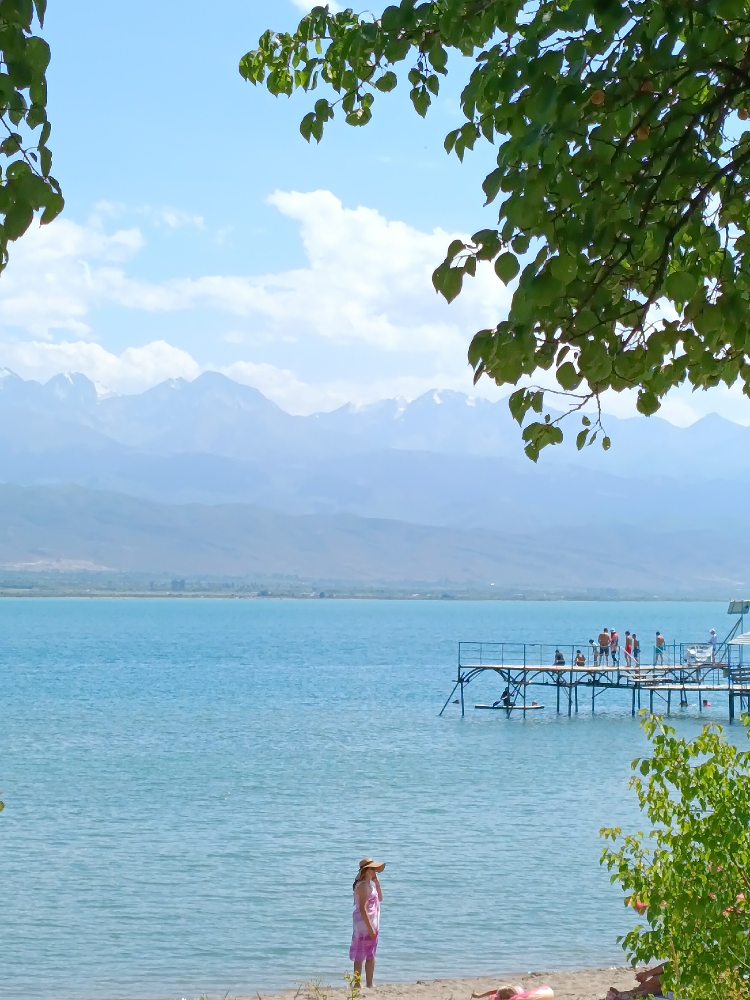 A wide shot captures a serene lake scene with distant, snow-capped mountains under a clear blue sky with scattered white clouds. In the foreground, lush green leaves frame the top of the image, suggesting a tree overhead. A sandy beach runs along the bottom, with a solitary woman wearing a straw hat and a pink dress standing near the water's edge. Further out on the lake, a wooden pier extends from the right side, populated by a group of people, some of whom are standing and others appear to be sitting or interacting. A person is visible on a paddleboard in the water near the pier, and another person is seen swimming. The water itself is a calm, bright blue. The overall impression is a tranquil summer day at a lakeside resort, likely in the vicinity of Kirpichny, Kyrgyzstan, given the mountainous backdrop.