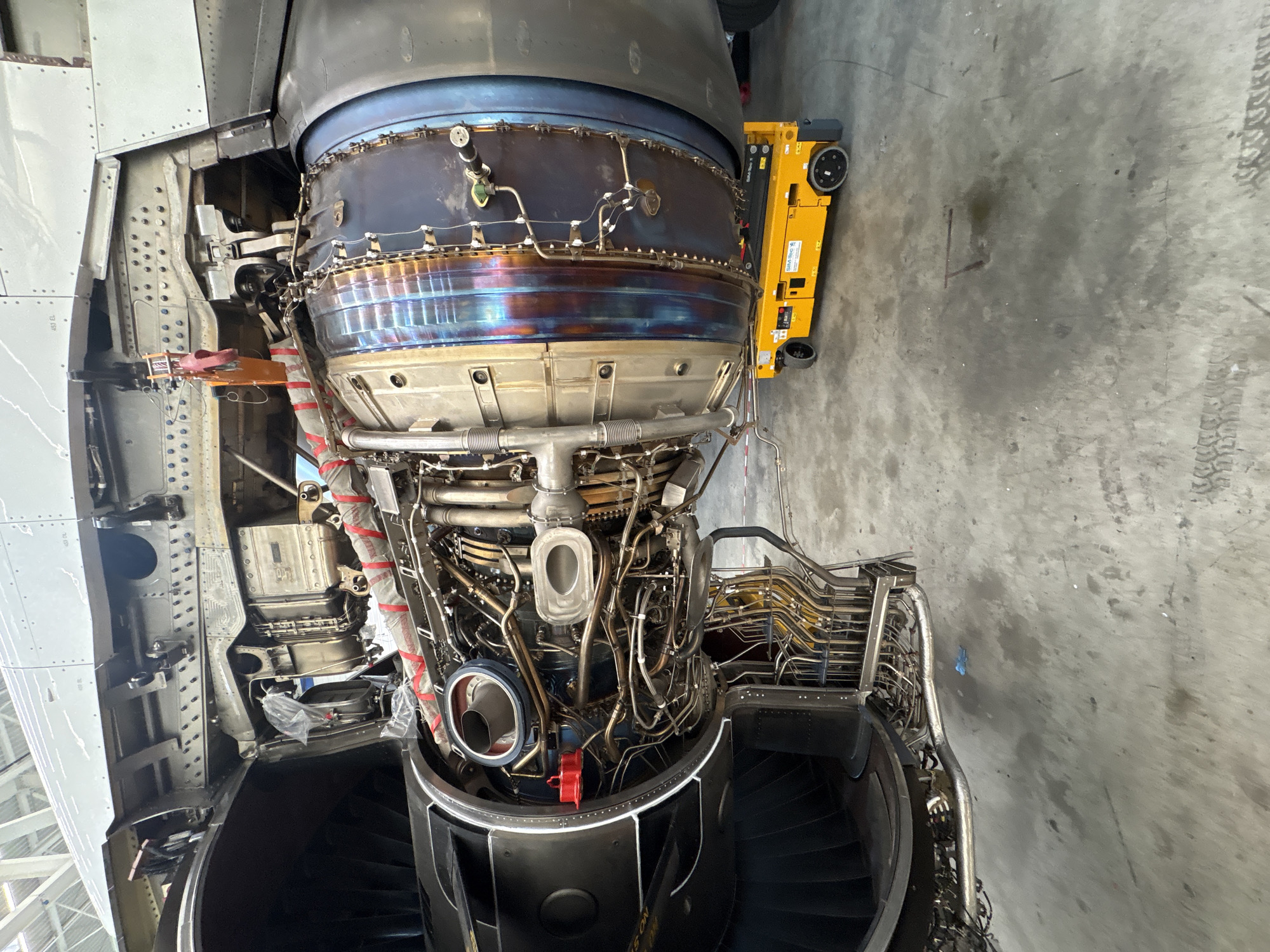 The image shows a disassembled aircraft engine, possibly undergoing maintenance or repair, inside a hangar or workshop. The engine is intricate with visible turbines, piping, and wiring. A yellow aircraft tow tractor is positioned adjacent to the engine. The location in the city of Lautzenhausen, Germany, suggests this is likely at or near the Frankfurt-Hahn Airport (HHN), which is nearby.