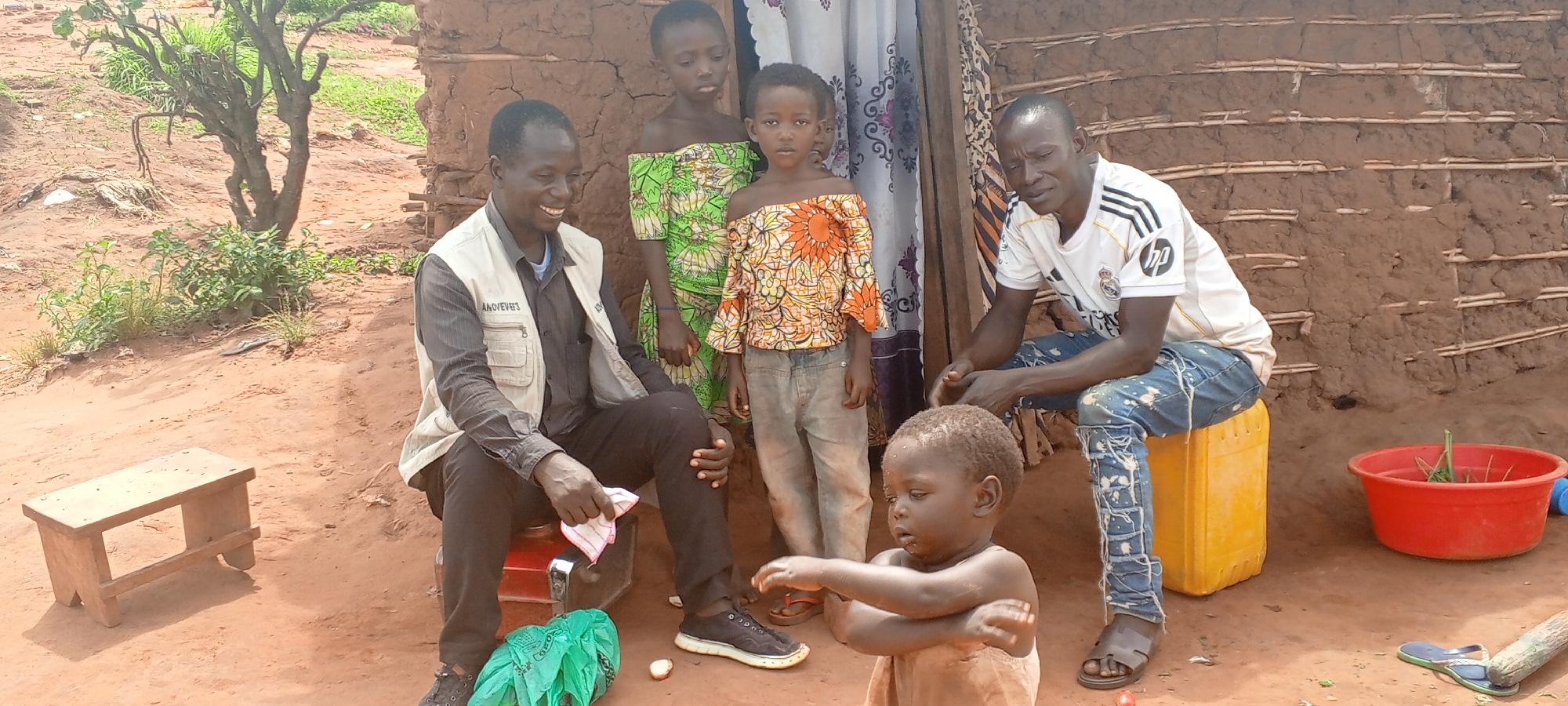 The scene depicts a group of individuals in what appears to be a rural setting in Makungu, Democratic Republic of the Congo. The backdrop consists of a mud-brick structure, possibly a dwelling. Five individuals are visible: two adults and three children. One adult, dressed in a light vest over a dark shirt and dark pants, is seated on what looks like a red container. He is holding a white cloth and appears to be smiling. The other adult is seated on a yellow container, wearing a white t-shirt and denim jeans, with sandals on his feet. A small child is standing in the foreground, wearing a simple garment. Behind them, two more children stand, one in a patterned green dress and the other in an off-the-shoulder orange and white top with grey pants. In the background, there's a small wooden stool and a red plastic container filled with greenery. The ground is primarily reddish-brown dirt. A small tree is visible on the left of the scene.