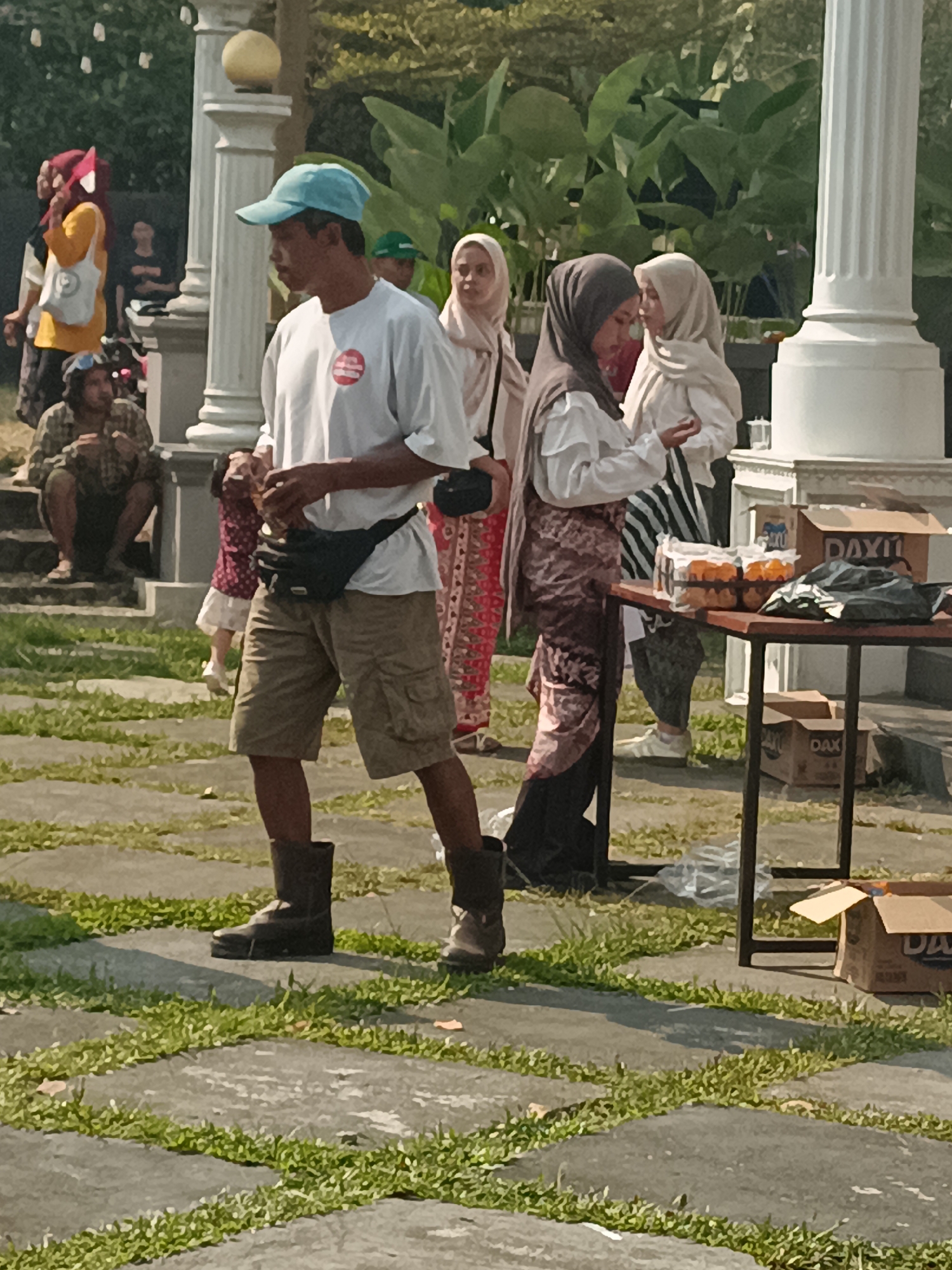 In Sleman, Indonesia, this image captures a bustling outdoor scene likely at a public gathering or market. The foreground is dominated by a man wearing a white t-shirt, khaki shorts, and tall brown boots. He has a black fanny pack around his waist and a light blue baseball cap on his head. The man is looking to his right, his hands clasped in front of him. Behind him, several individuals are visible. To his left, a person in a yellow top and red headscarf stands near a white column. Further left, a person sits on stone steps, appearing to be eating. To the man's right, two women in hijabs are interacting with each other, one wearing a light brown hijab and the other a darker brown one. They are standing near a table laden with food items, including what appears to be trays of cupcakes. Boxes are stacked on and around the table. The background features lush green foliage and more columns, suggesting a park or an area with architectural features. The ground is paved with stone slabs interspersed with patches of grass, indicating an outdoor public space. The lighting suggests it is daytime, likely morning or afternoon.