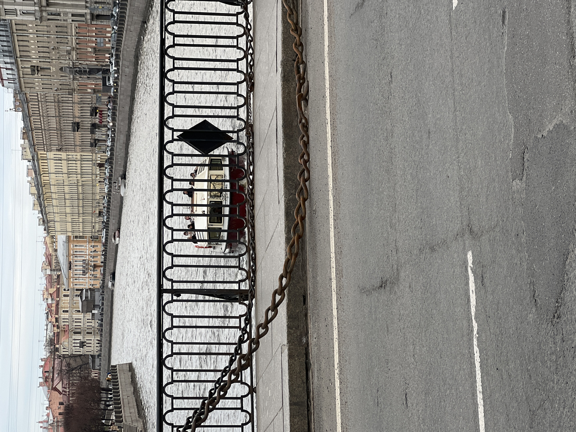 The image captures a canal scene in Saint Petersburg, Russia, under an overcast sky. In the foreground, a grey asphalt road runs parallel to the canal, marked by a faded white painted line. A heavy, rusty metal chain is draped along the pavement next to the road. A black metal fence, featuring ornate, repeating oval and bar patterns with a diamond-shaped accent, separates the pavement from the canal.
Beyond the fence, the calm, grey water of the canal fills the middle ground. A white and red boat, possibly a tour vessel, is visible in the canal, with a few small figures of people on board. The far bank of the canal is lined with a continuous array of historic, multi-story buildings, characterized by numerous windows and facades in various shades of beige, grey, and light yellow, typical of European architecture. Further along the bank, smaller structures and sparse, bare trees are visible.
The sky is uniformly light grey, indicating an overcast daytime. The overall atmosphere is calm and subdued. No specific events are actively taking place, and there is no visible text.