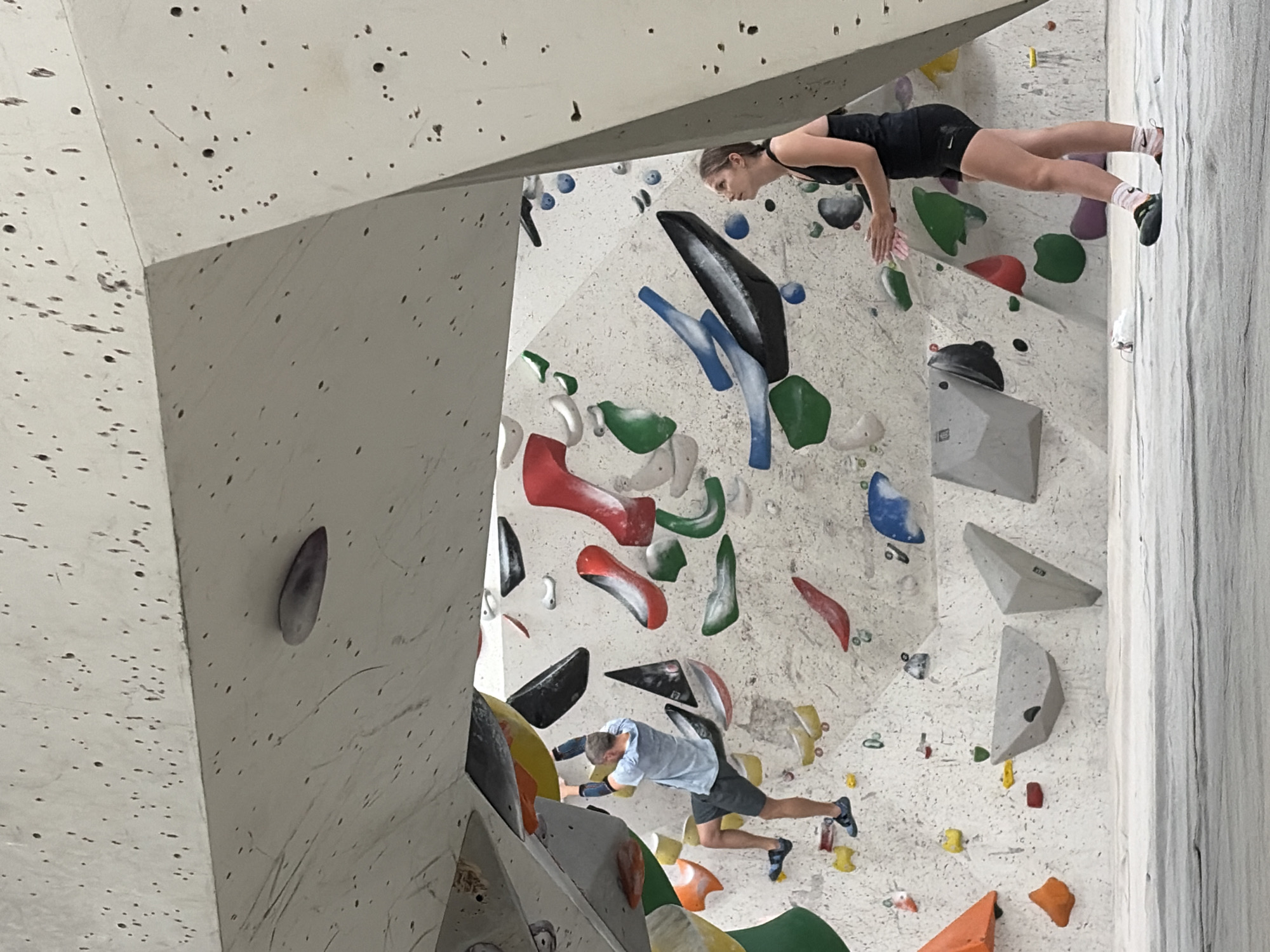This is a medium shot, full-color, eye-level photograph taken inside a climbing gym. The gym features a large, white, angular wall with various climbing holds in bright colors. A man in a light blue t-shirt and dark shorts is climbing on the left side of the wall, reaching for a yellow hold. He is wearing climbing shoes and arm guards. On the right side of the frame, a woman in a black tank top and shorts stands on the mat below, looking up at the man. She is wearing athletic socks and climbing shoes. The floor is covered in a white, padded mat. The background consists of the textured, concrete climbing wall with numerous colorful holds. The lighting appears to be artificial, casting soft shadows. The image depicts a scene of recreational rock climbing. The location is likely a climbing gym in Khimki, Russia.