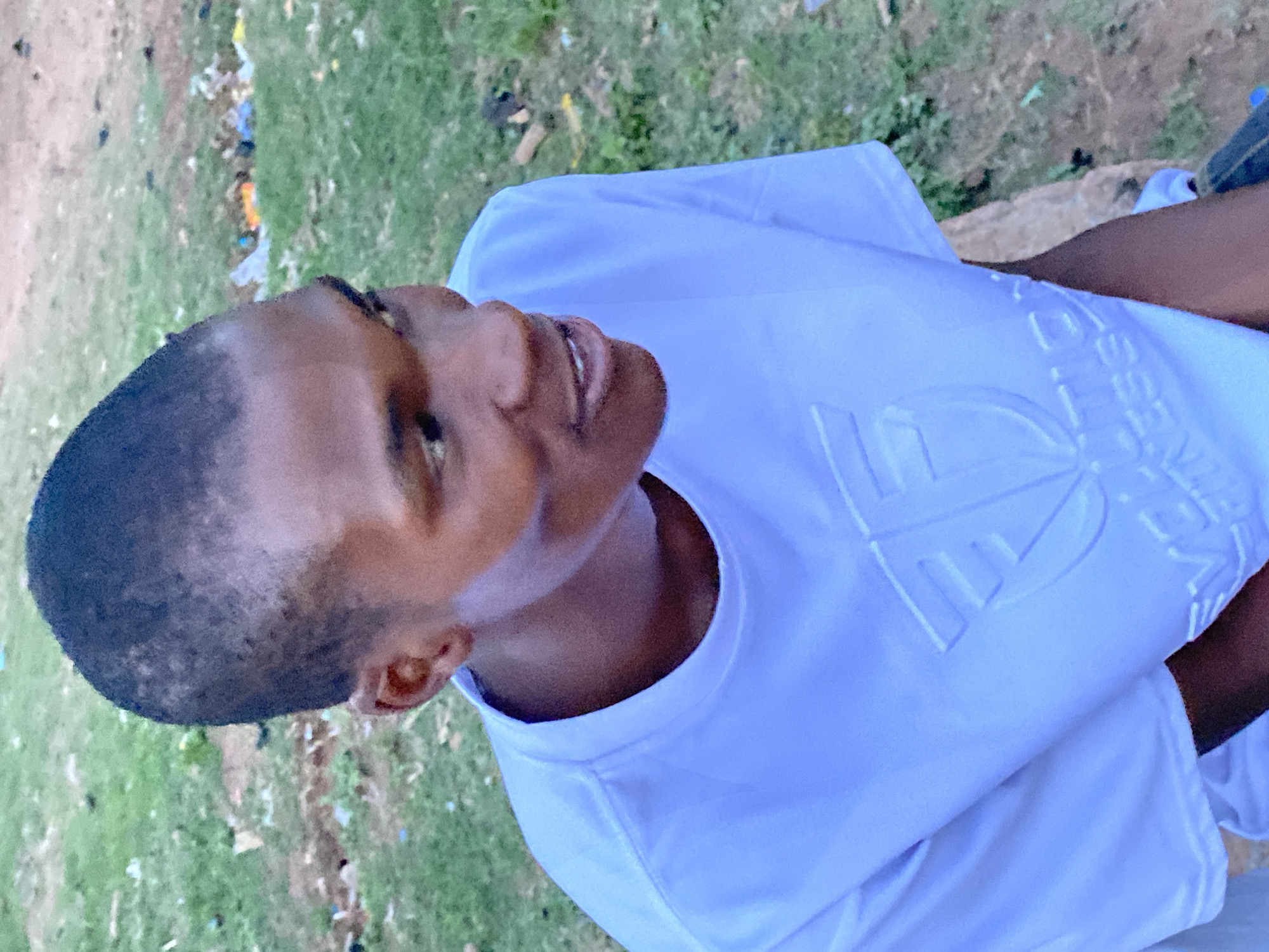 A young man with dark hair and skin, wearing a white t-shirt with embossed lettering, stands in an outdoor location in Bukuru, Nigeria. The surrounding environment features grass and possibly some debris on the ground. The photo is taken from a low angle, looking upwards at the subject, who appears to be glancing slightly upwards and to the side.