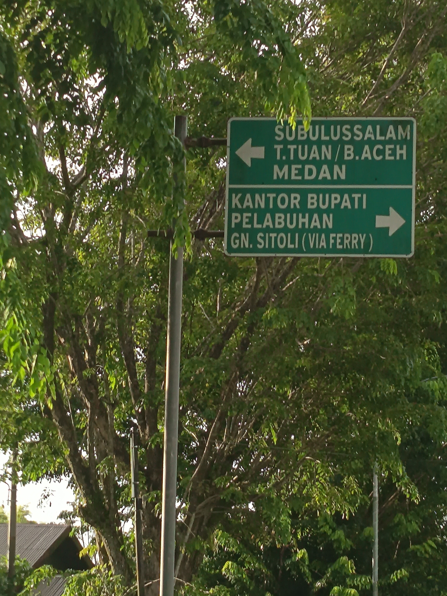 A directional sign is pictured in the city of Singkil, Indonesia. The sign has two green rectangular panels with white text and arrows. The top panel indicates "SUBULUSSALAM T.TUAN/B.ACEH MEDAN" with an arrow pointing left. The bottom panel reads "KANTOR BUPATI PELABUHAN GN. SITOLI (VIA FERRY)" with an arrow pointing right. The sign is mounted on a metal pole. Behind the sign, lush green trees with dense foliage dominate the background. Part of a building's roof is visible in the lower left corner.